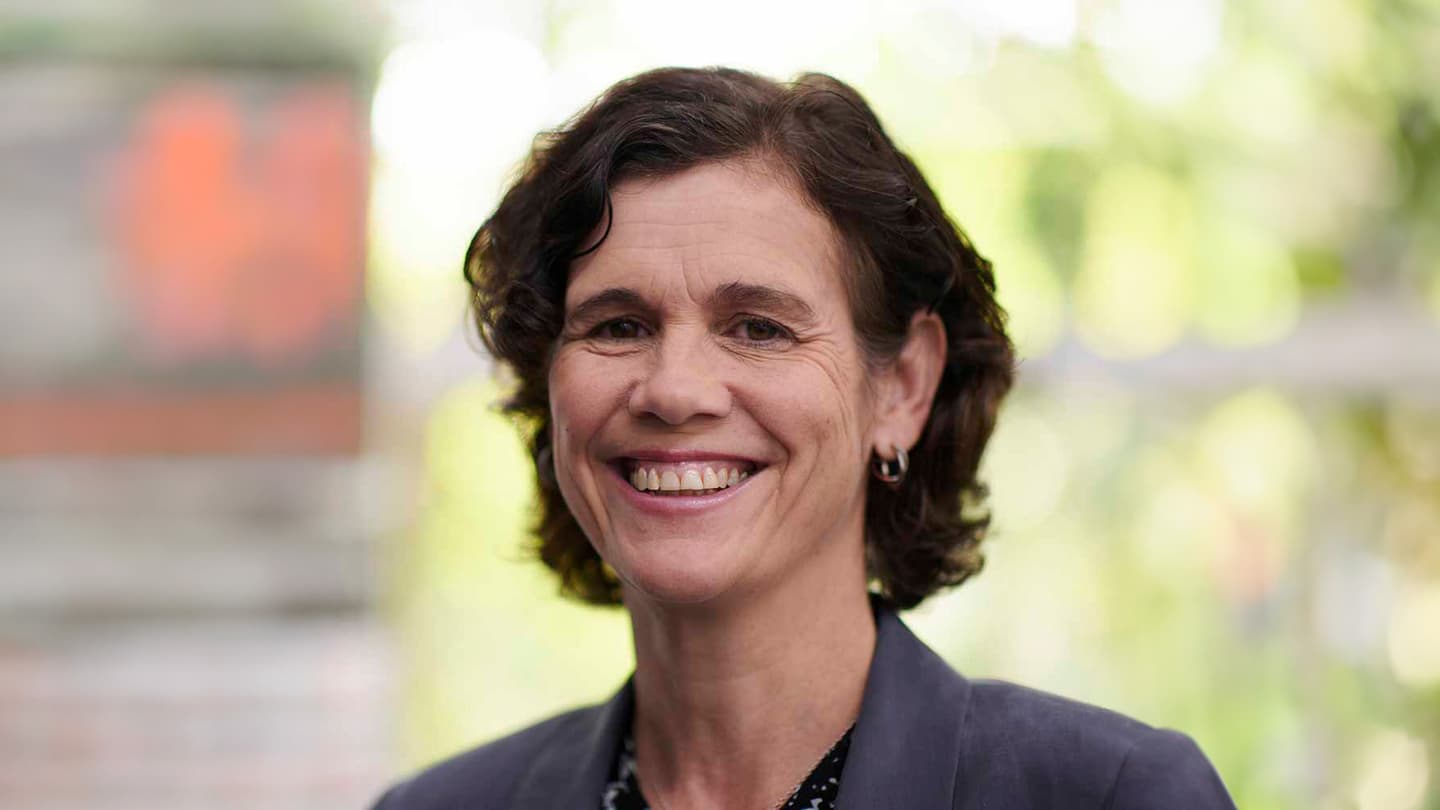 Person with short wavy dark hair smiling broadly, wearing a dark blazer against a soft-focus green background.