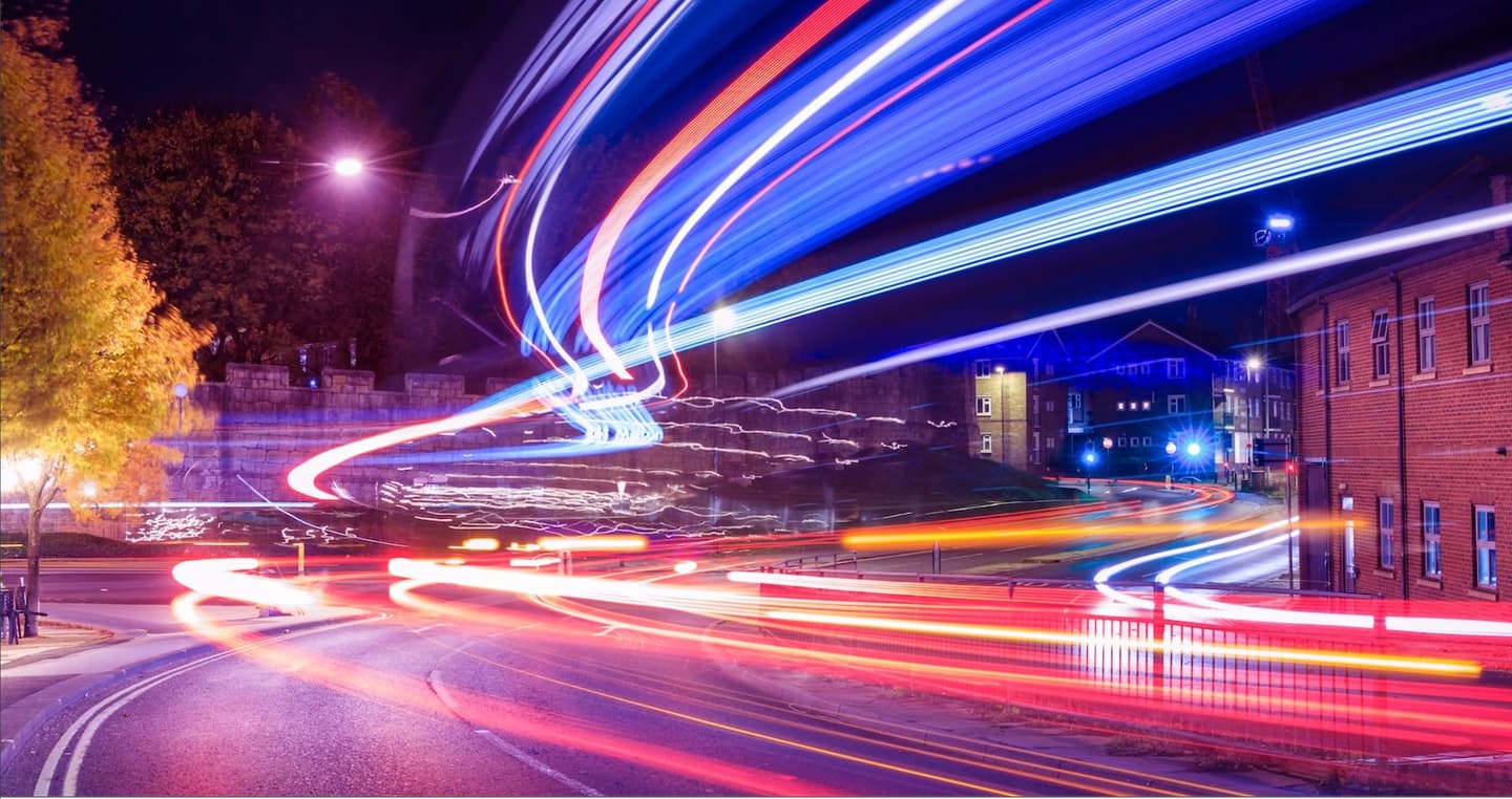 Long exposure night photograph showing colourful light trails from traffic against urban buildings and autumn trees.