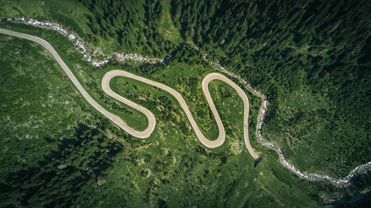 Aerial view of a winding mountain road with sharp hairpin bends cutting through lush green forest and rocky terrain.