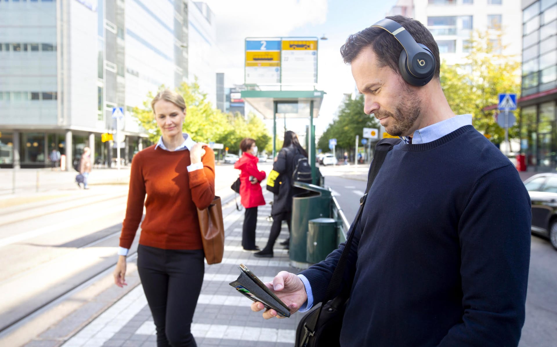 Person wearing headphones checking phone at bus stop in city, with commuters and urban buildings in background.