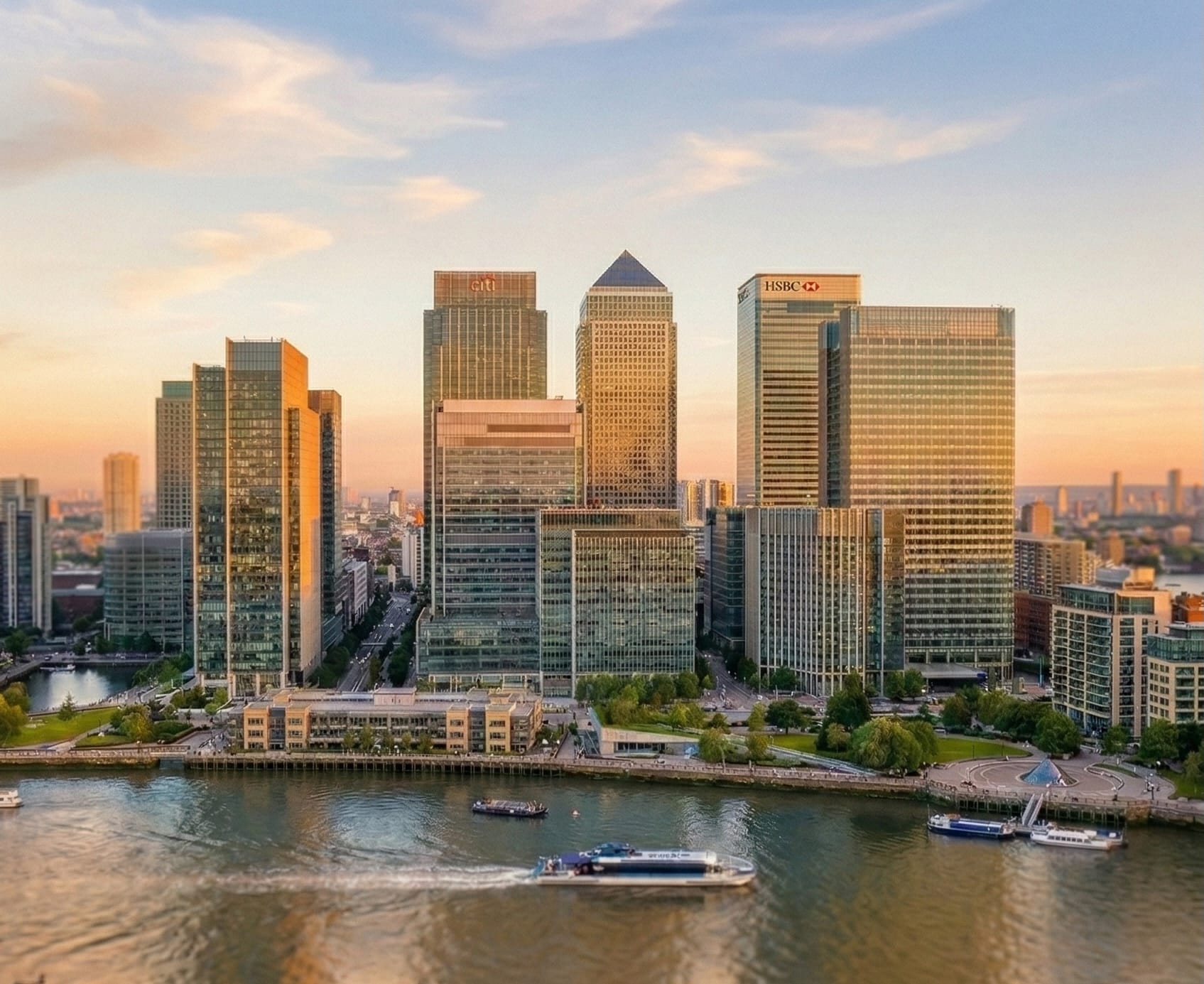 Canary Wharf skyline at sunset with Citi and HSBC towers reflecting golden light, boats cruising on the Thames below.