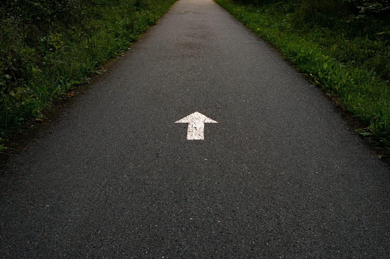 A narrow tarmac path stretching into the distance, with a white forward arrow painted on it, flanked by lush green vegetation.