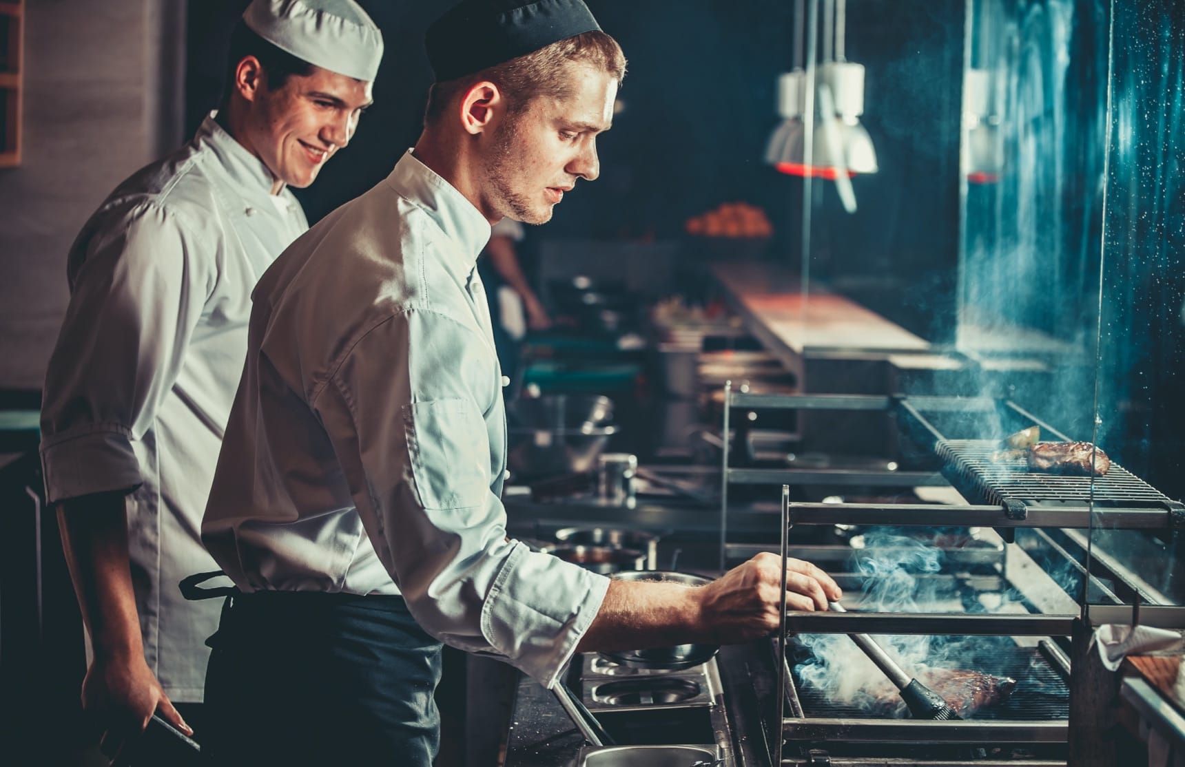 Two chefs in white uniforms working at a grill station in a dimly lit restaurant kitchen with rising smoke.