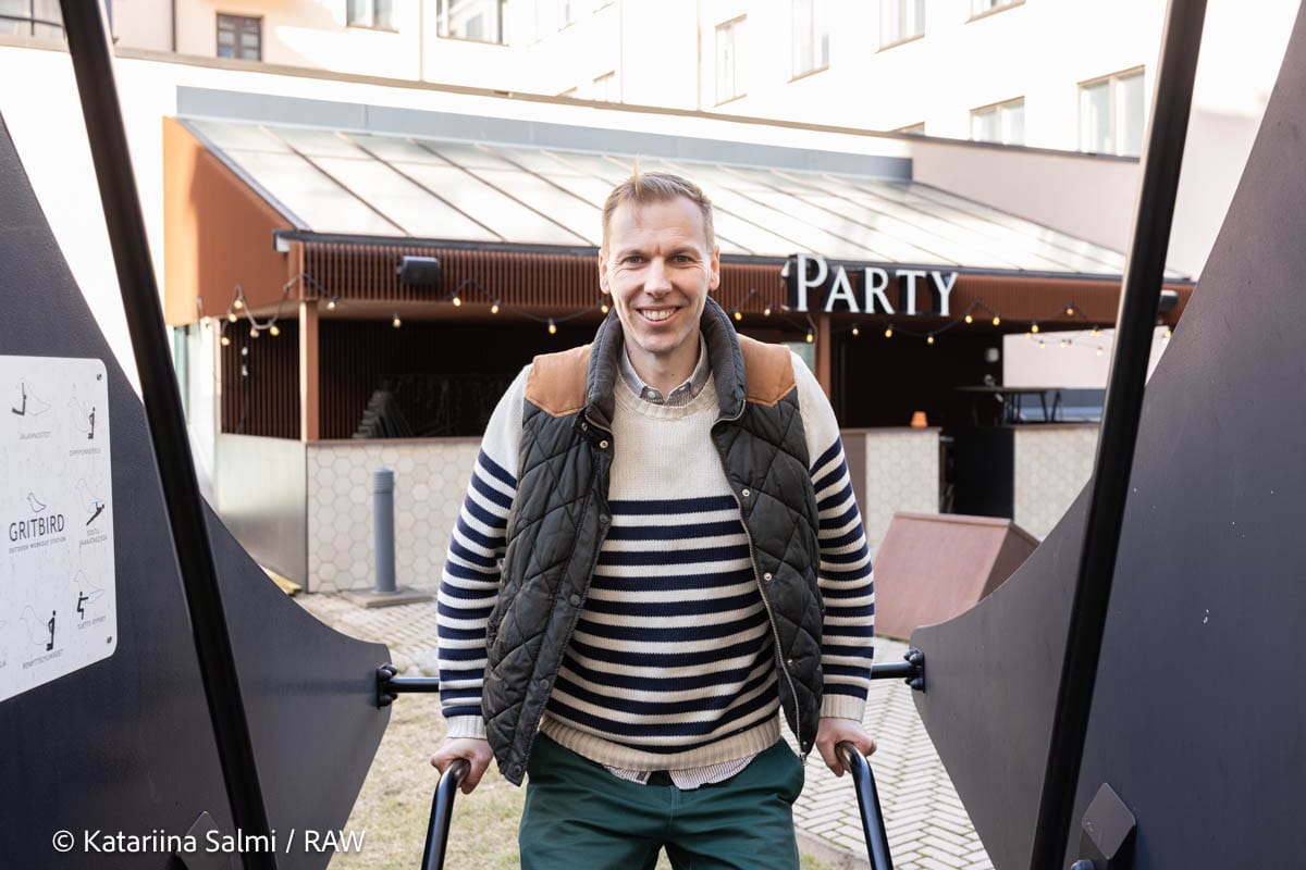 Person in striped jumper and quilted gilet smiling outside a restaurant with "Party" signage visible.