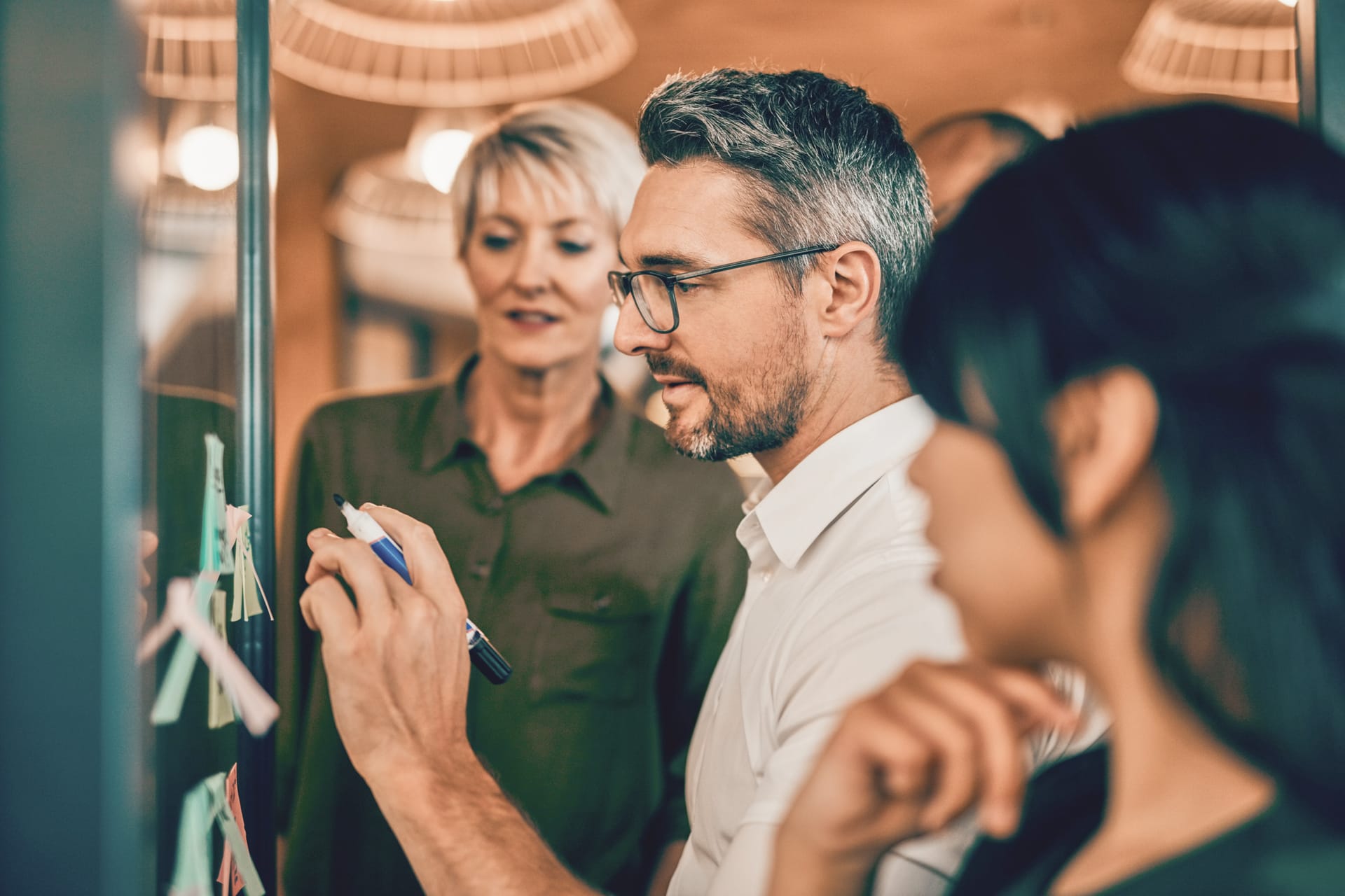 Two colleagues in an office setting reviewing notes on a glass board, with warm lighting and pendant lamps above.