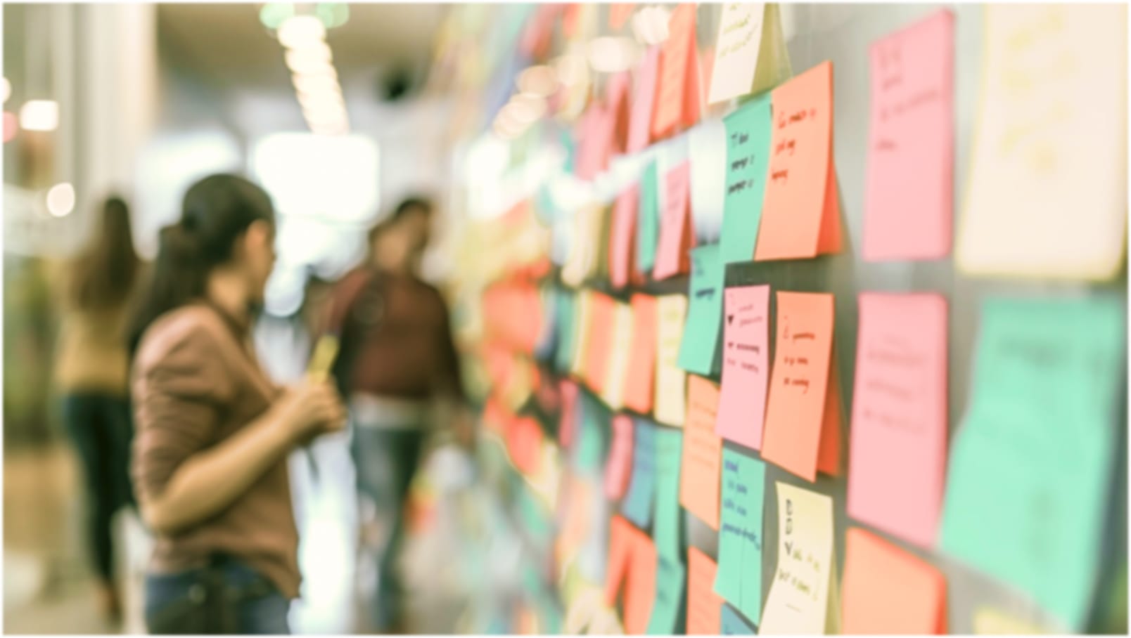 A board covered in colourful sticky notes with blurred people discussing ideas in the background.