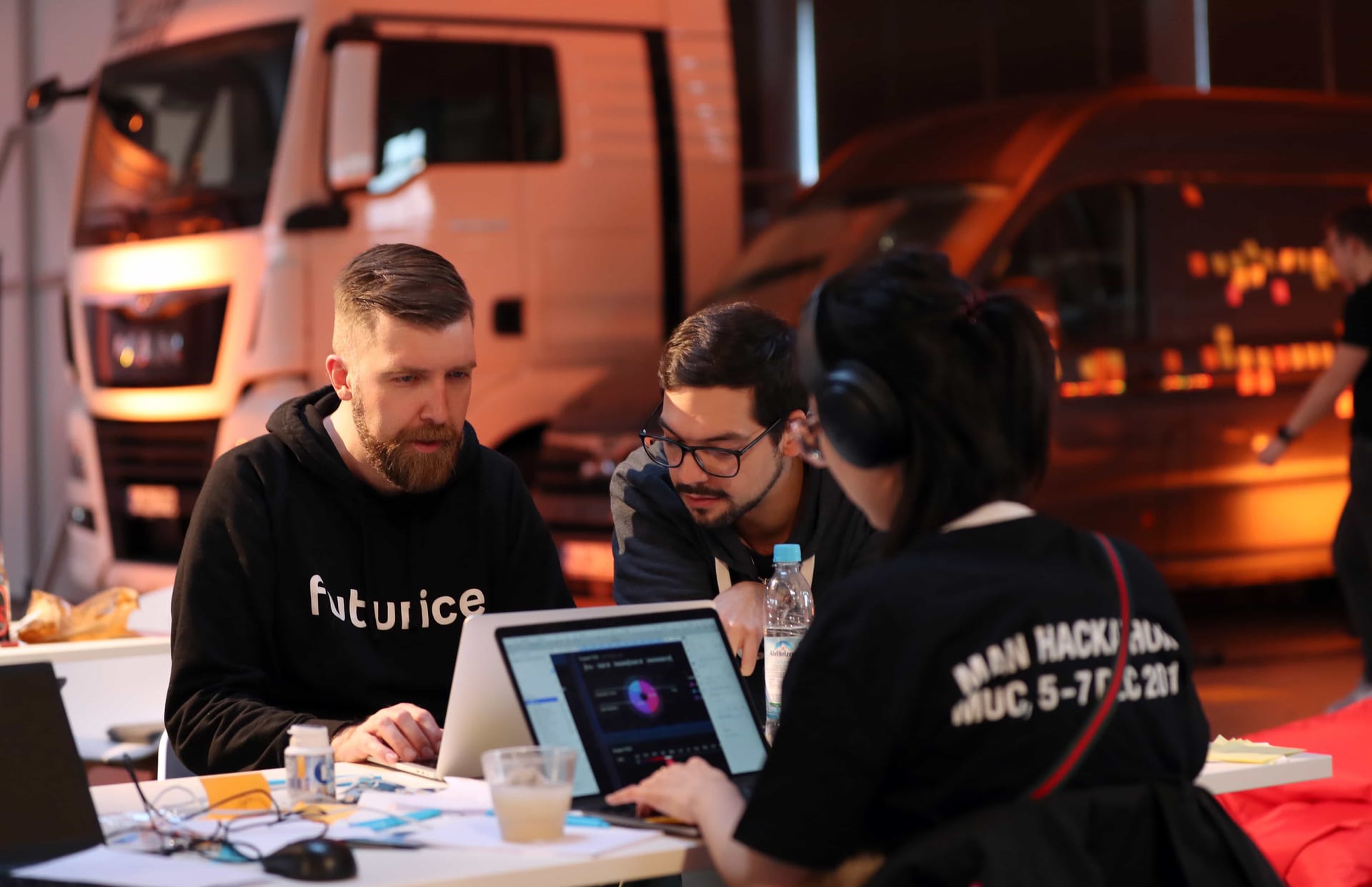 Team collaborating on laptop at hackathon event with trucks visible in background, warm evening lighting.