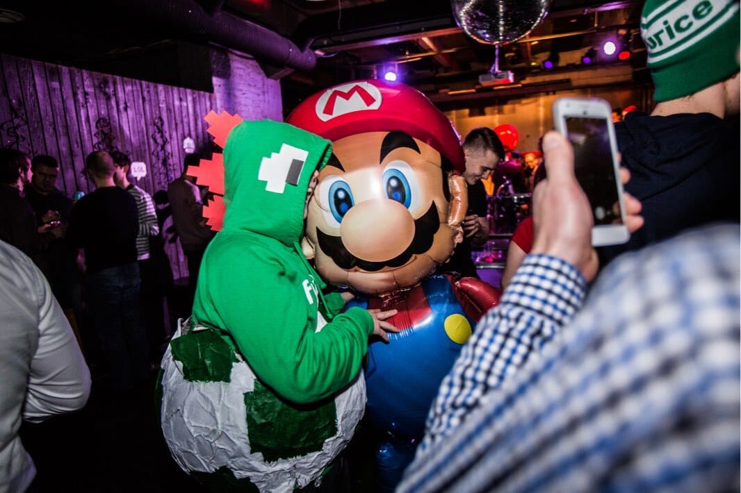Person in Yoshi costume posing with Mario balloon at nightclub party with disco ball and colourful lighting.