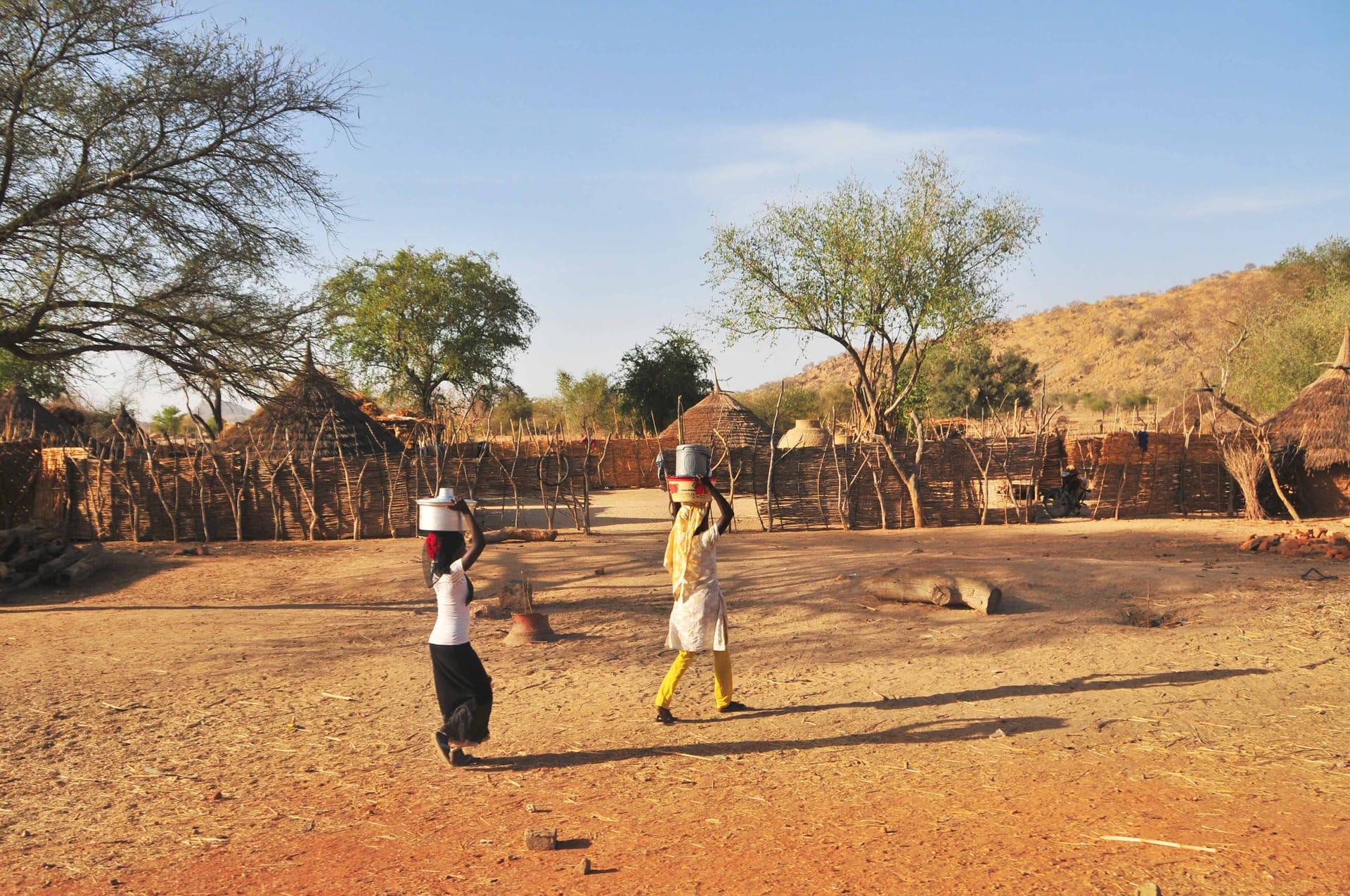 Two people carrying water containers on their heads walking through an African village with thatched huts and wooden fences.
