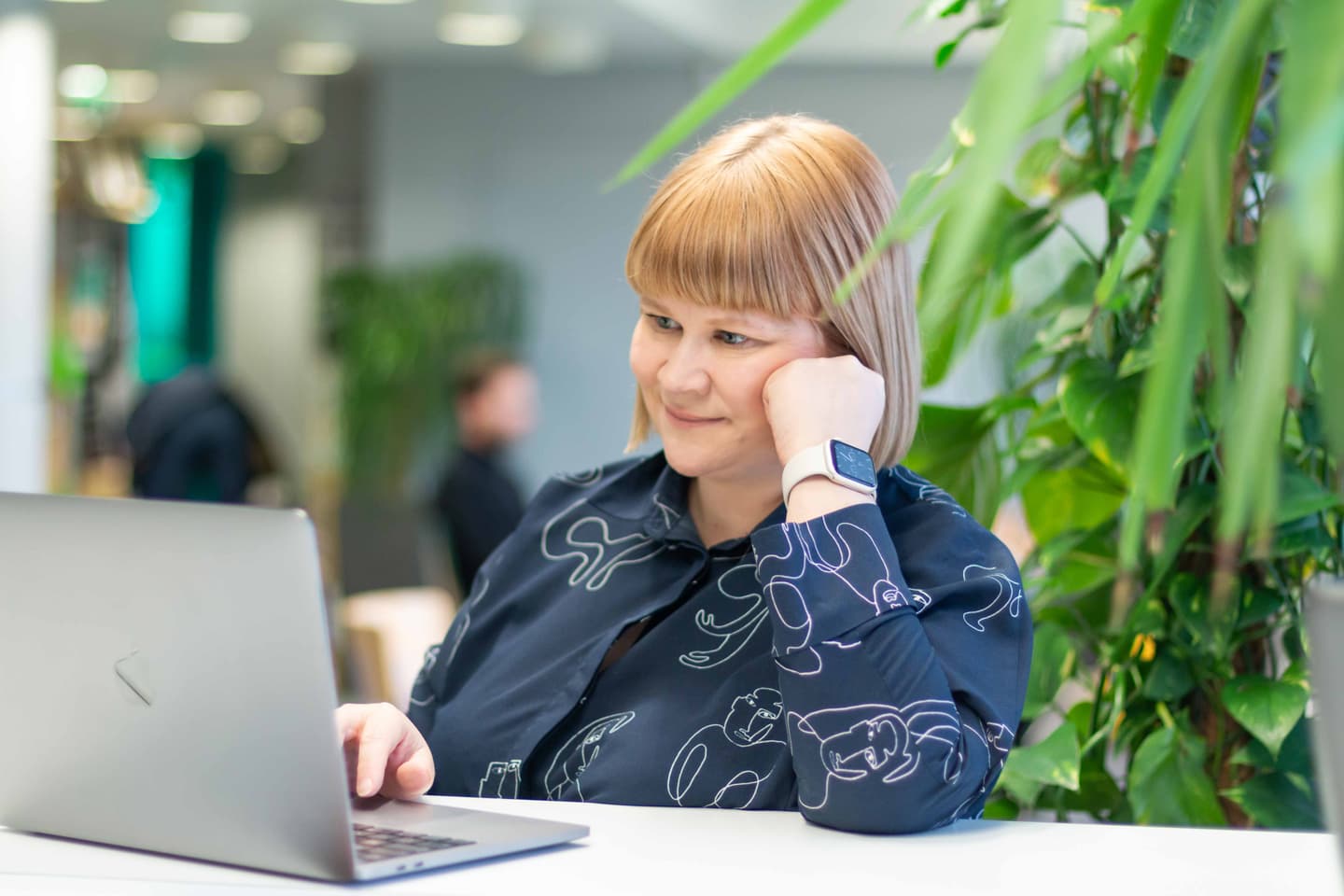Person with blonde hair wearing a patterned blue shirt and smartwatch working on laptop in green plant-filled office space.