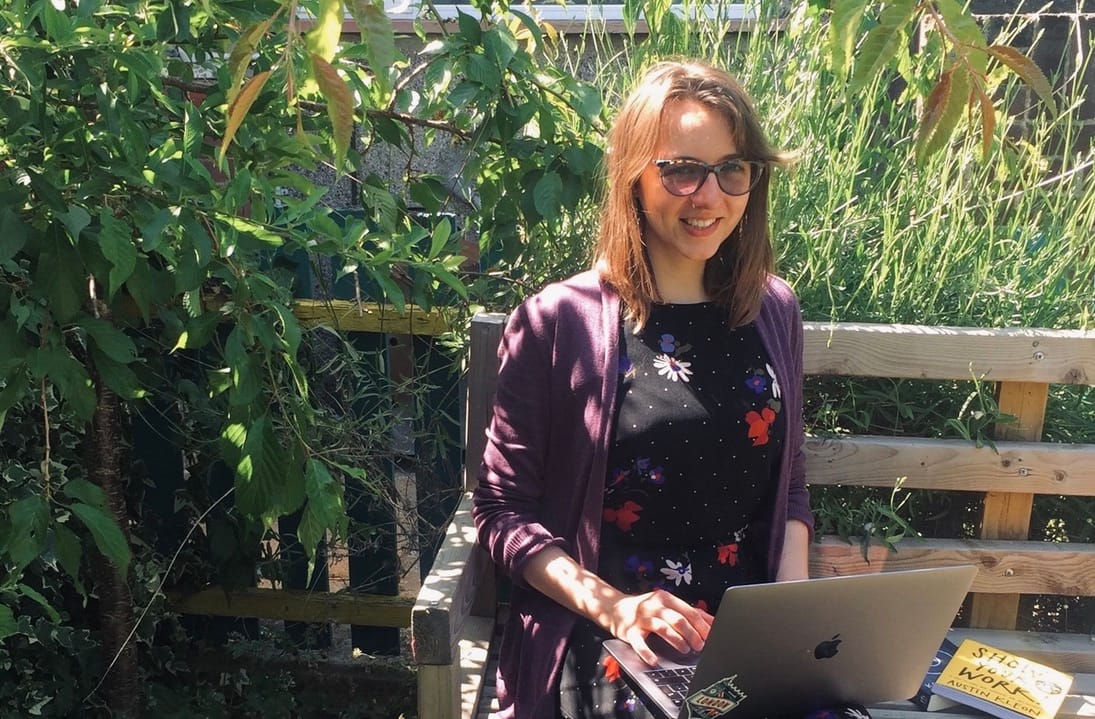Person in sunglasses working on laptop outdoors in a lush garden setting, wearing purple cardigan over floral top.
