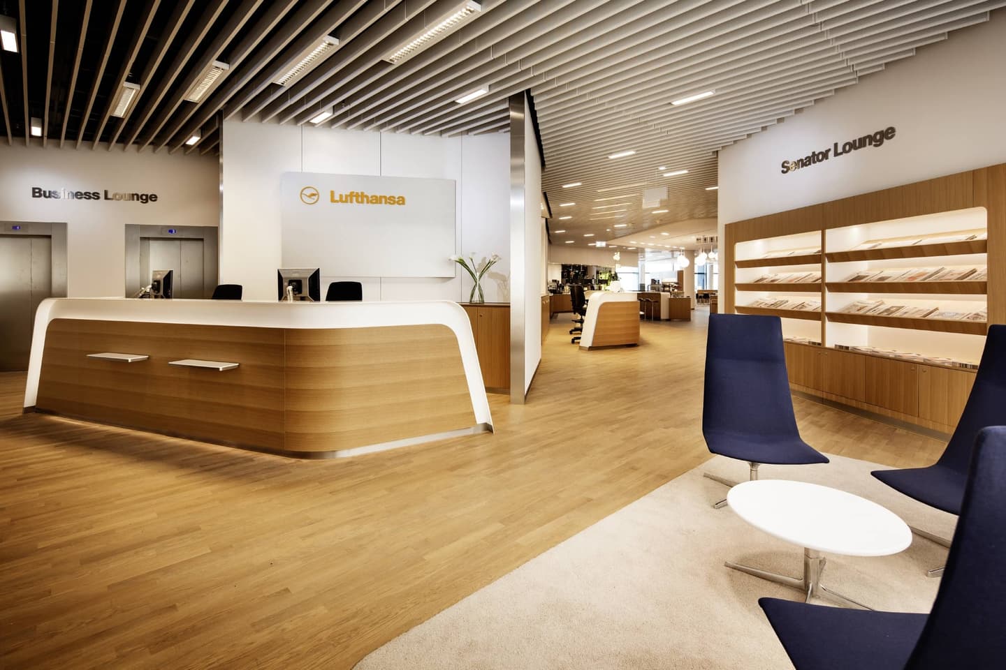 Modern Lufthansa airport lounge with wooden reception desk, blue chairs, and separate Business and Senator lounge areas.