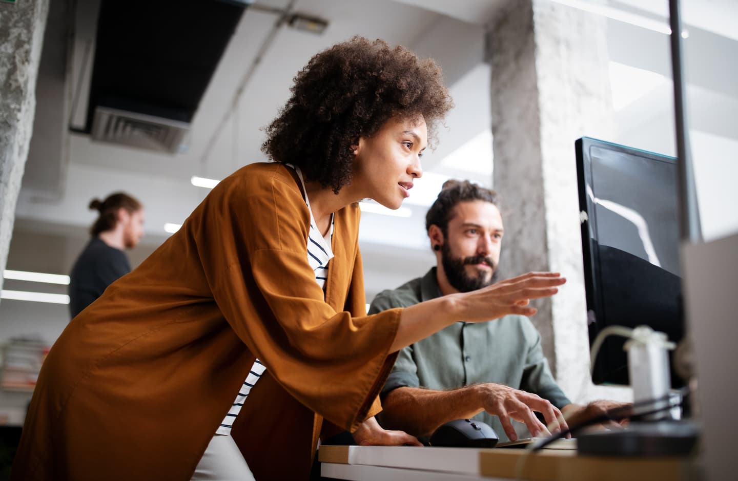 Person in brown jacket pointing at computer screen while colleague in grey shirt works at desk in modern office.