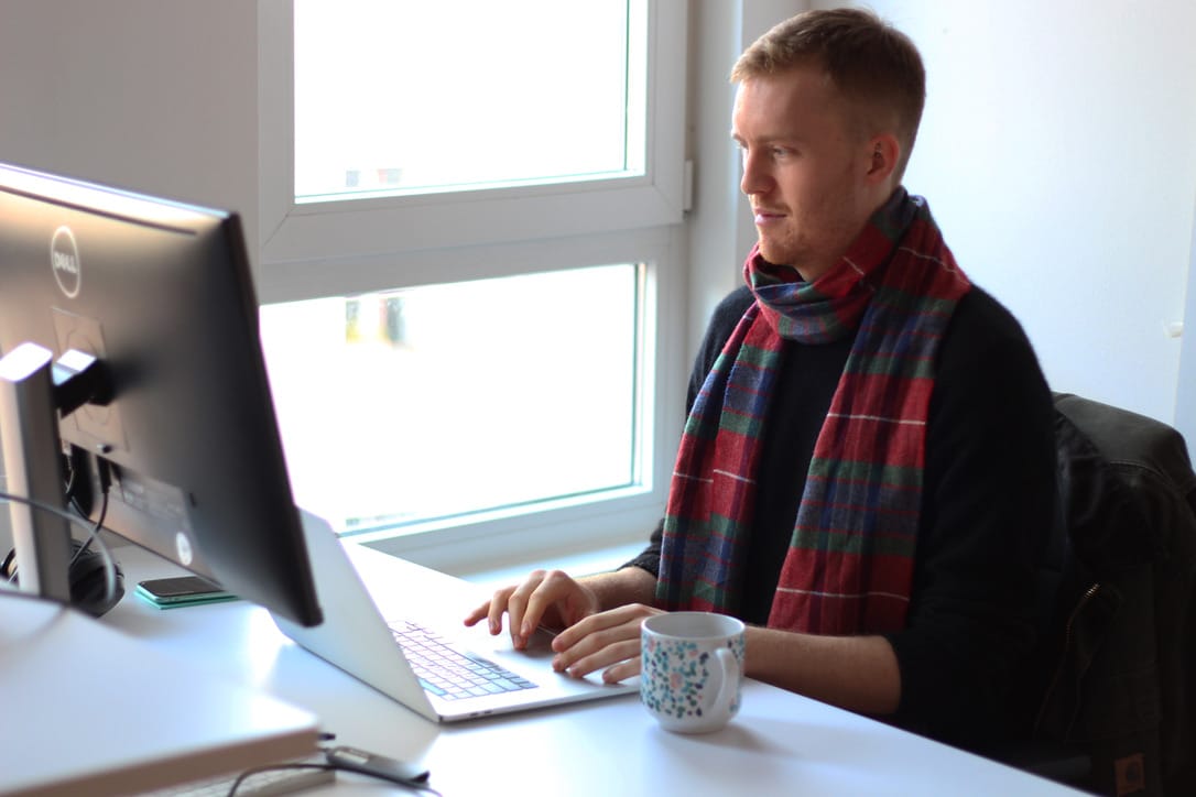 Person in plaid scarf working on laptop at desk with coffee mug, seated by bright window.