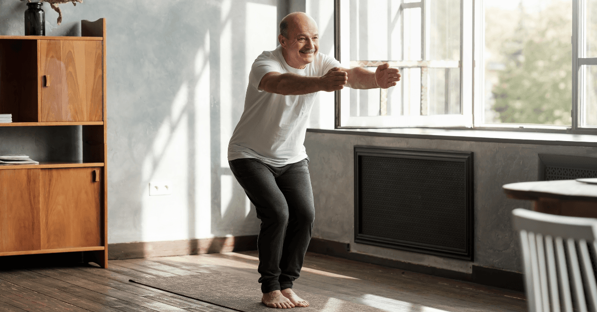 Older man in white t-shirt exercising in a bright room with wooden floors and furniture, smiling while stretching.