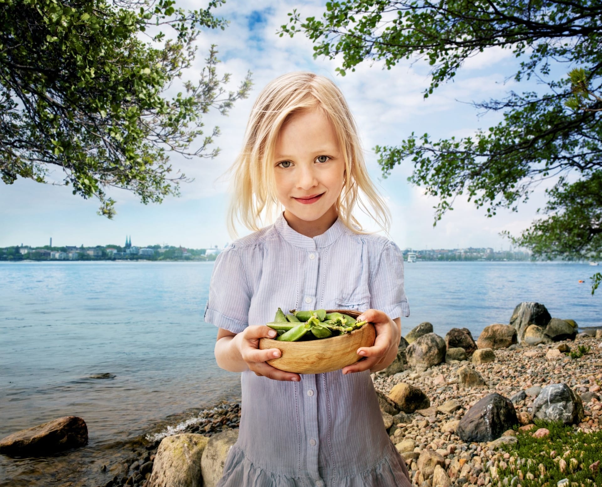 Child in light blue shirt holding wooden bowl of greens by lakeside with rocky shore and city skyline in background.