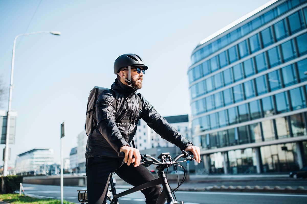 Cyclist in black jacket and helmet riding through urban area with modern glass office buildings against blue sky.