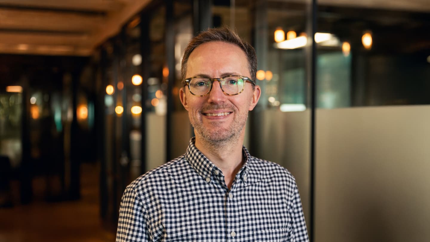 Matthew Billson of Piclo Energy in a checkered shirt, smiling ina  modern office space with warm lighting in the background.