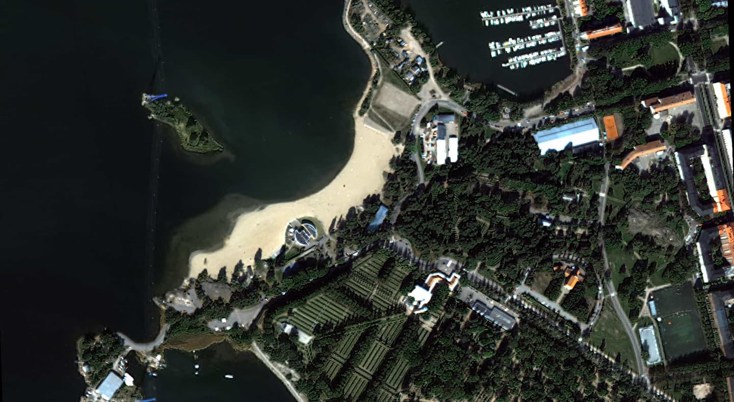 Aerial view of a coastal area with sandy beach, marina with boats, forested land, and organised agricultural fields nearby.