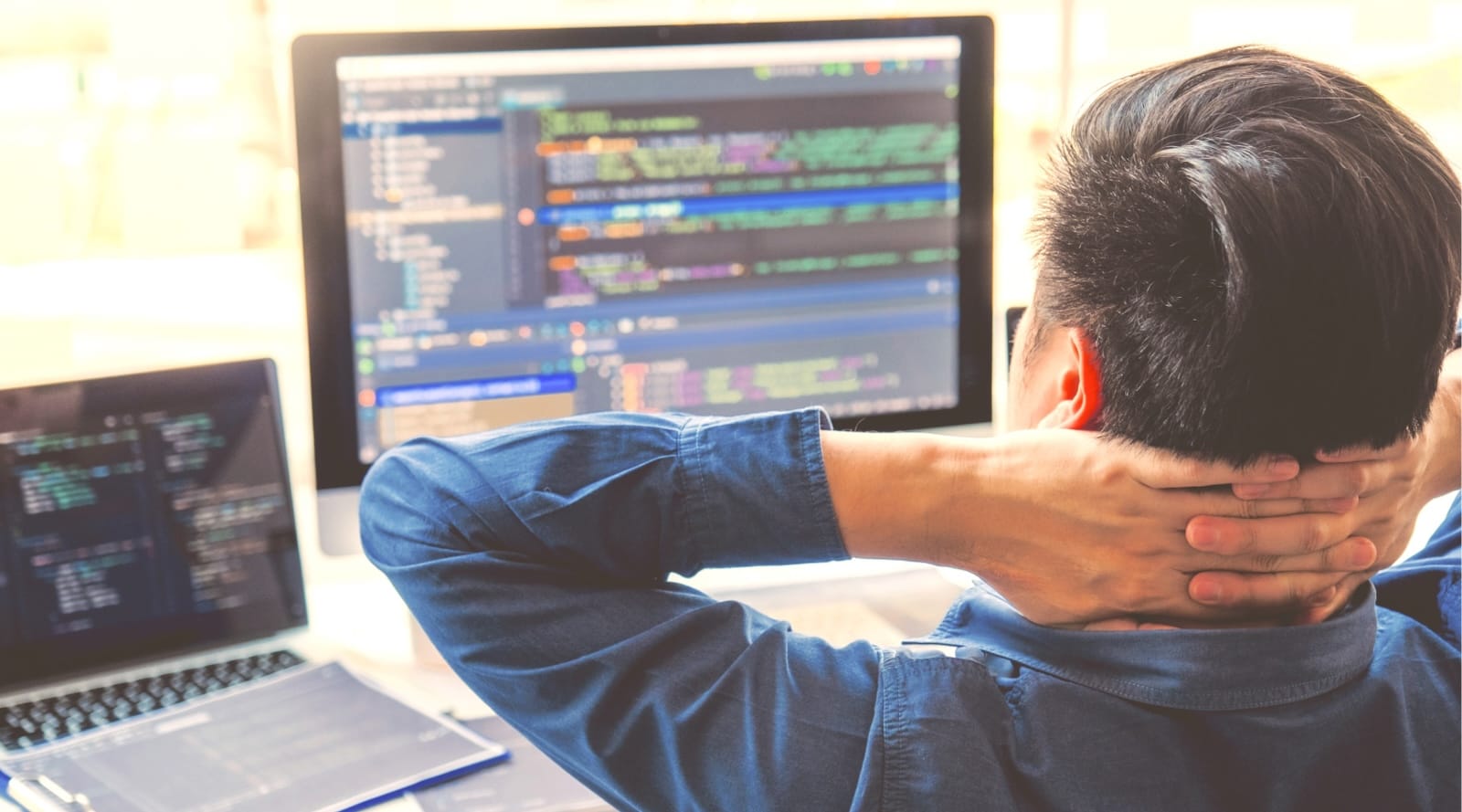 A man in a blue shirt sits with hands behind his neck, staring at monitors displaying colourful programming code.