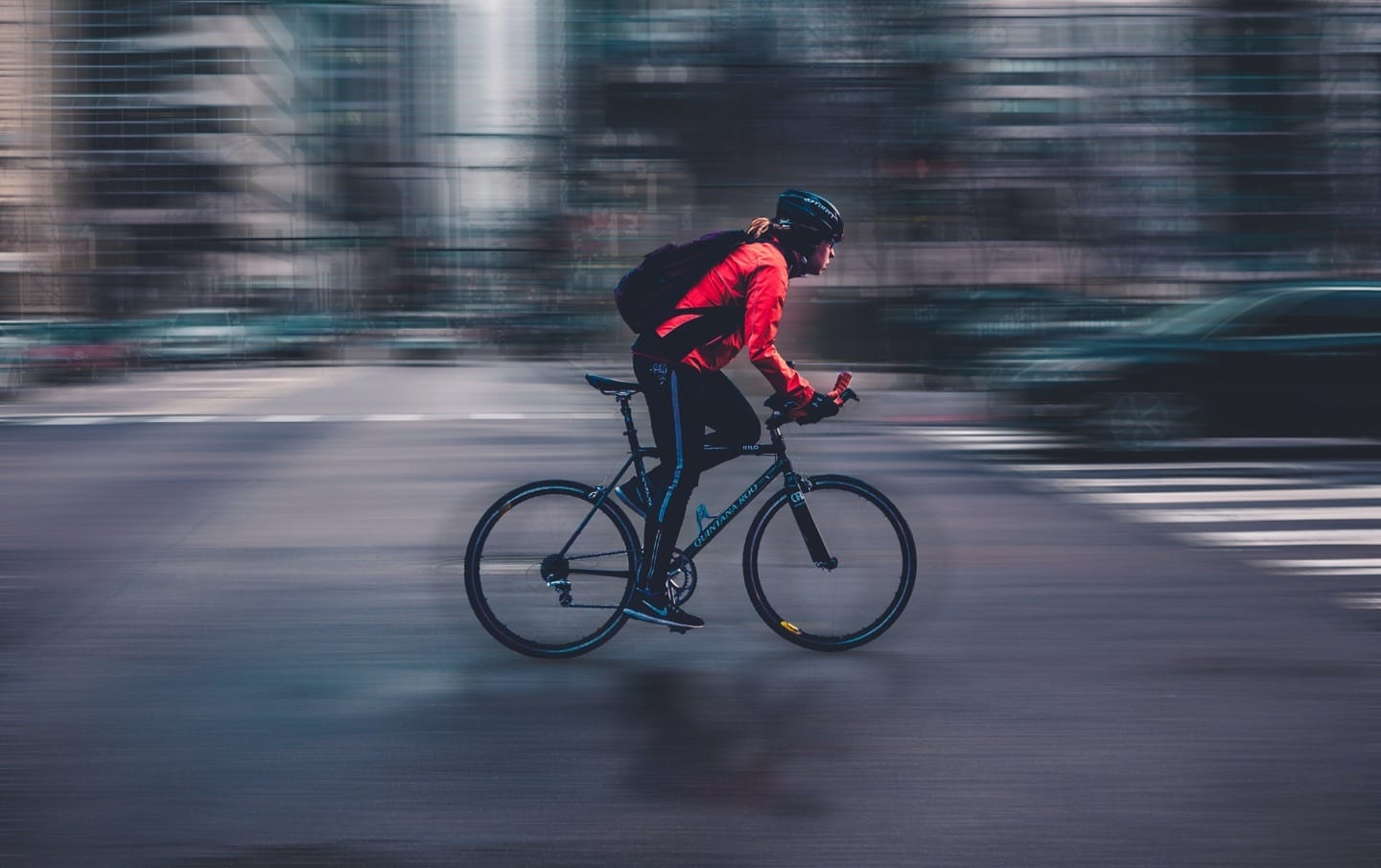 Cyclist in bright red jacket and helmet speeding through city streets with motion blur effect in the background.