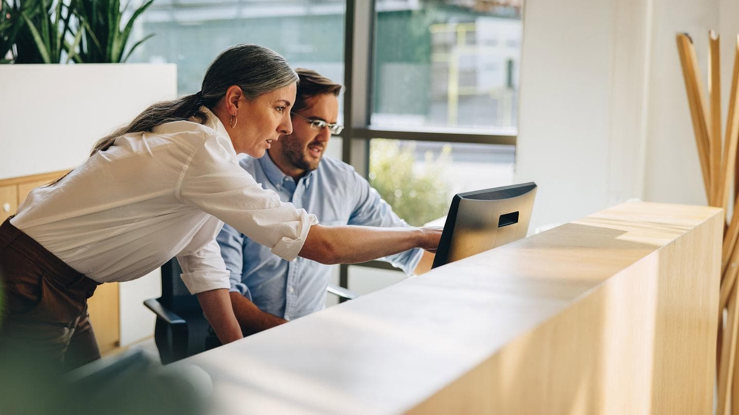 Two colleagues looking at a computer screen at a reception desk in a bright, modern office with large windows.