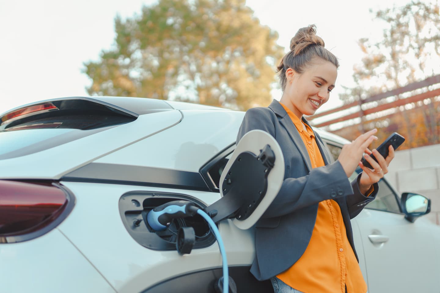 Person in blue blazer and orange shirt checking phone while charging white electric car outdoors in an autumn setting.