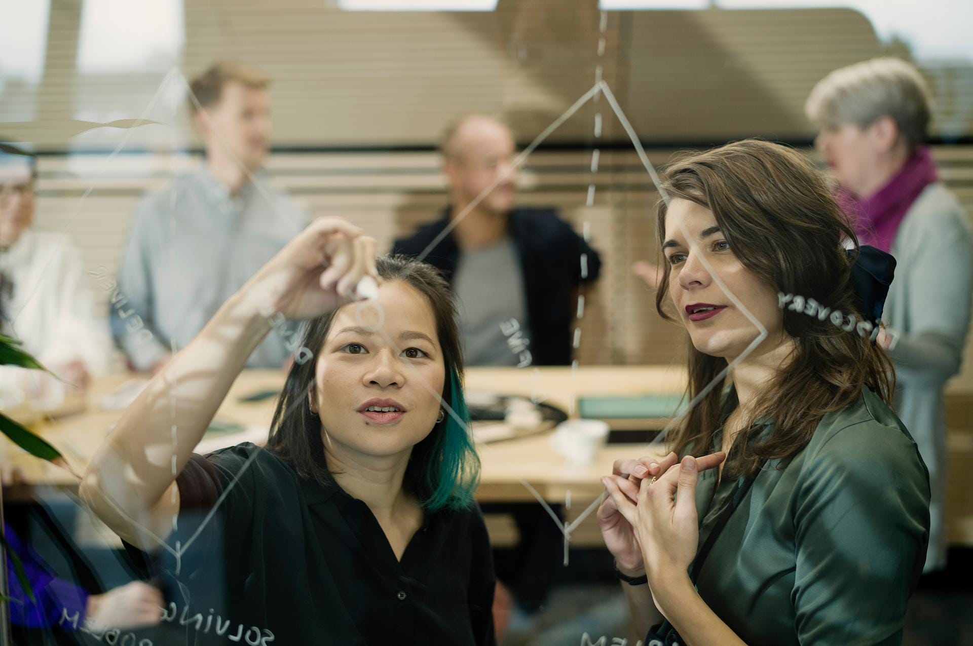 Two women collaborating at a glass board with colleagues visible in the background during a business meeting.