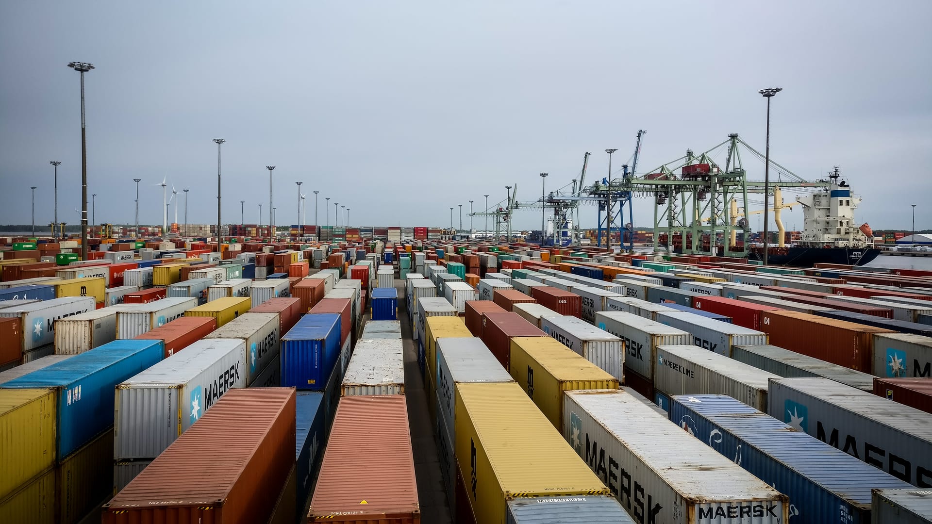 Busy shipping port with rows of colourful cargo containers, cranes and a docked vessel under an overcast sky.