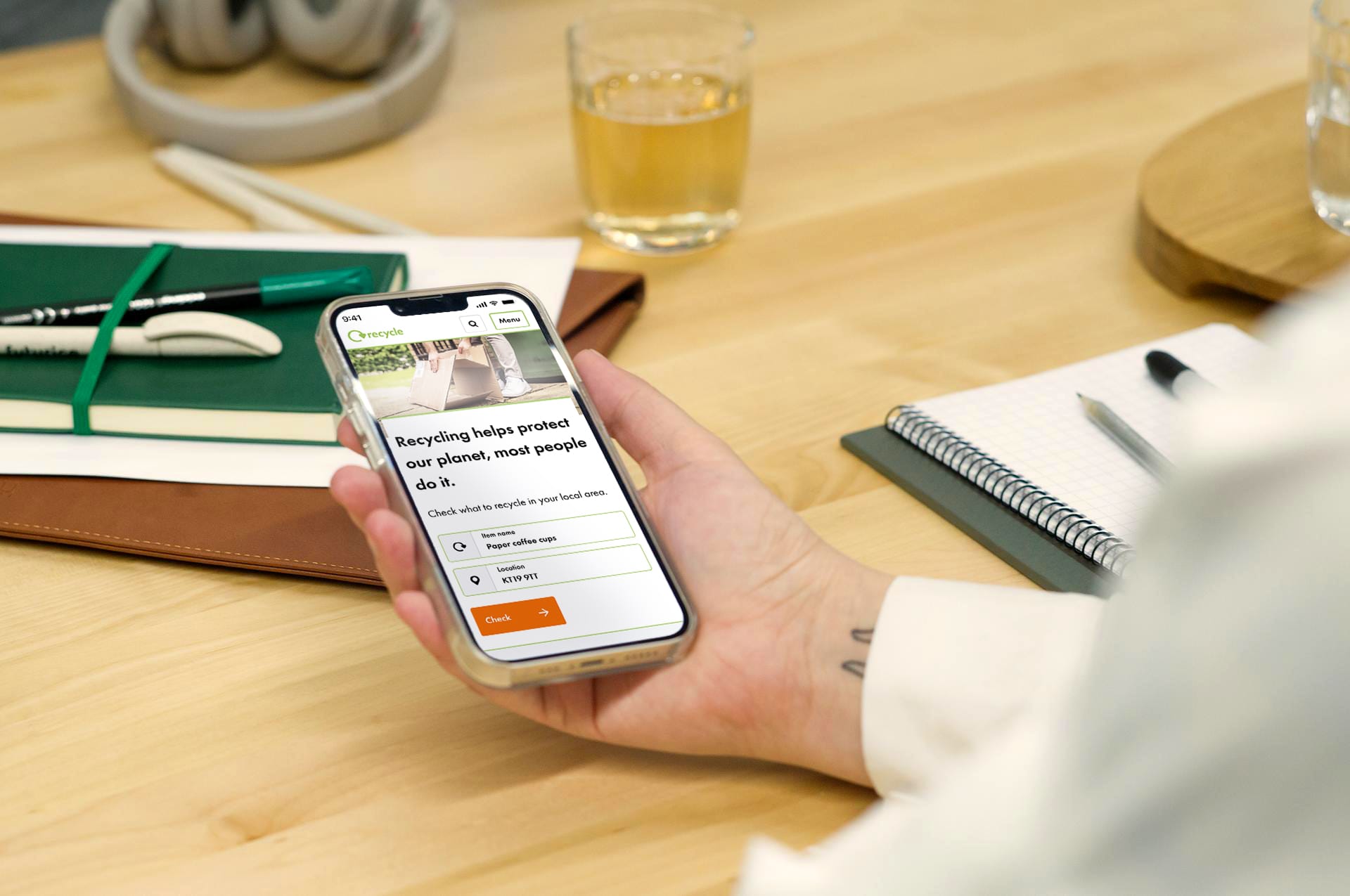 Hand holding phone displaying recycling app with search for paper coffee cups on wooden desk with notebook and drink.