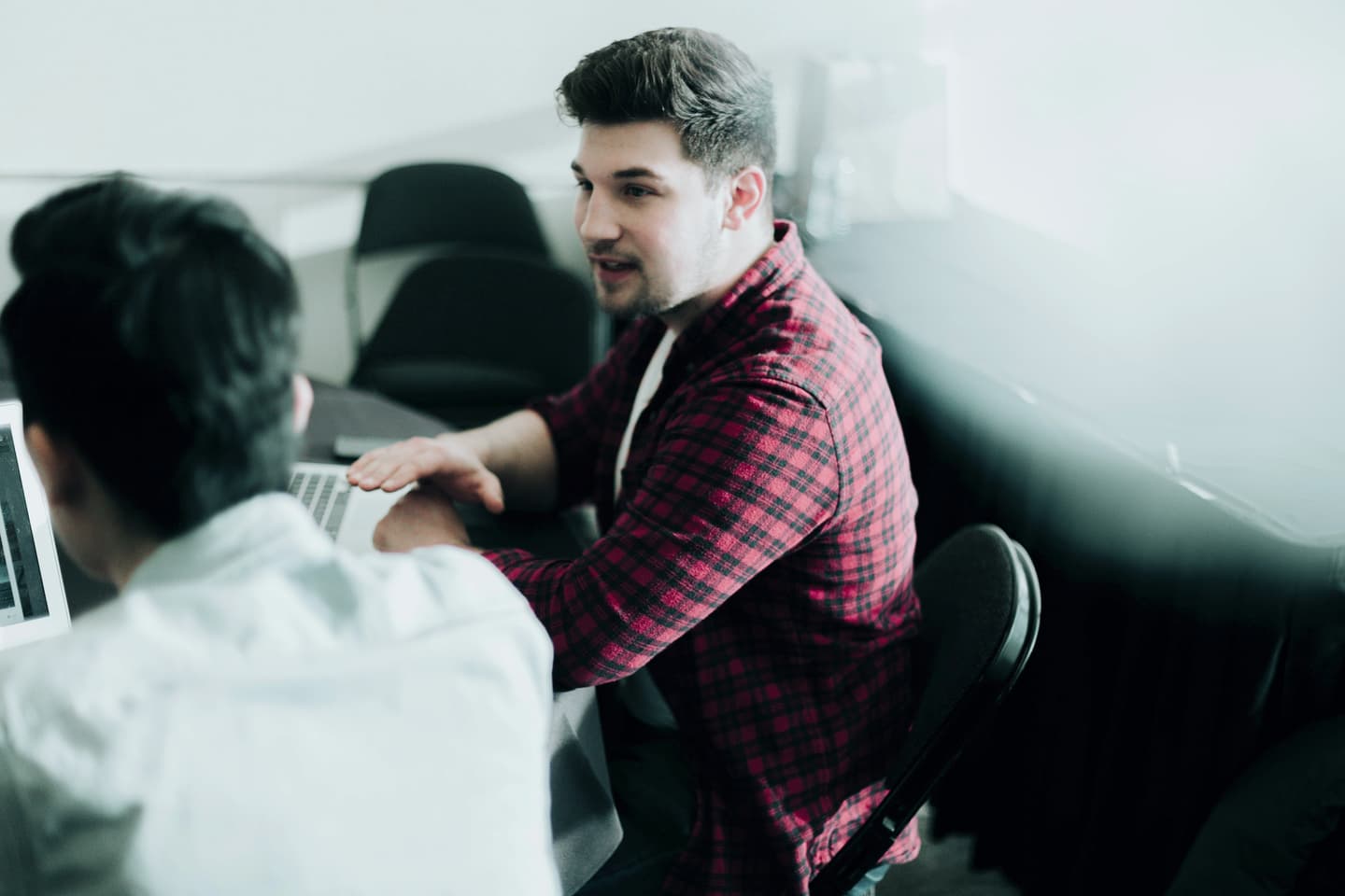 Person in red plaid shirt speaking with colleague in white shirt during office meeting, laptop visible on table.