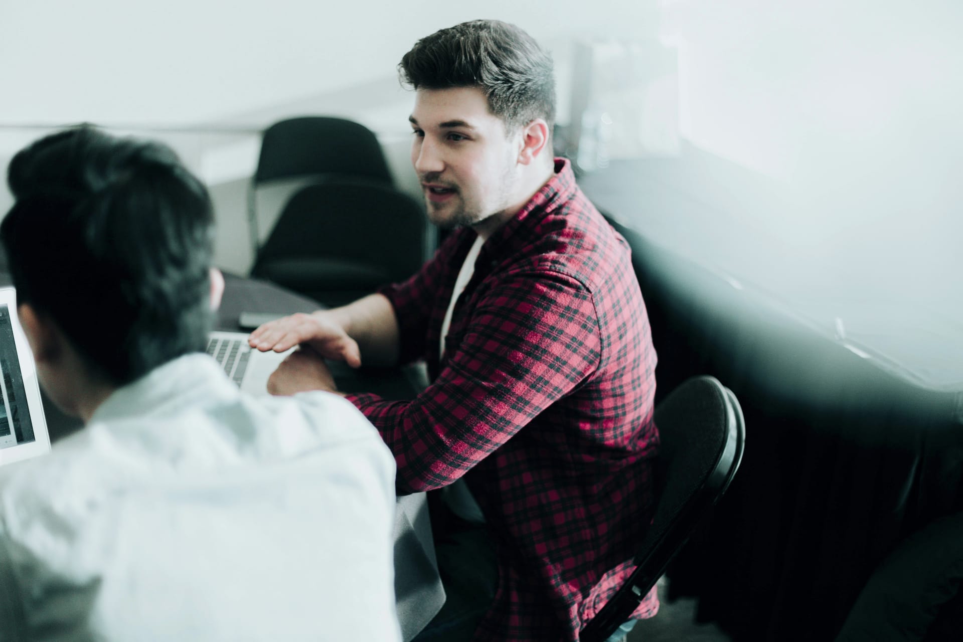 Person in red plaid shirt speaking with colleague in white shirt during office meeting, laptop visible on table.
