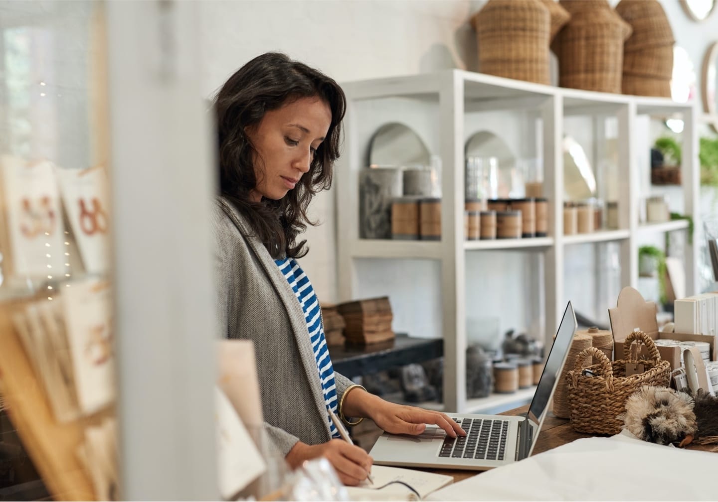 Person working on laptop in stylish retail shop with white shelving displaying natural products and woven baskets.