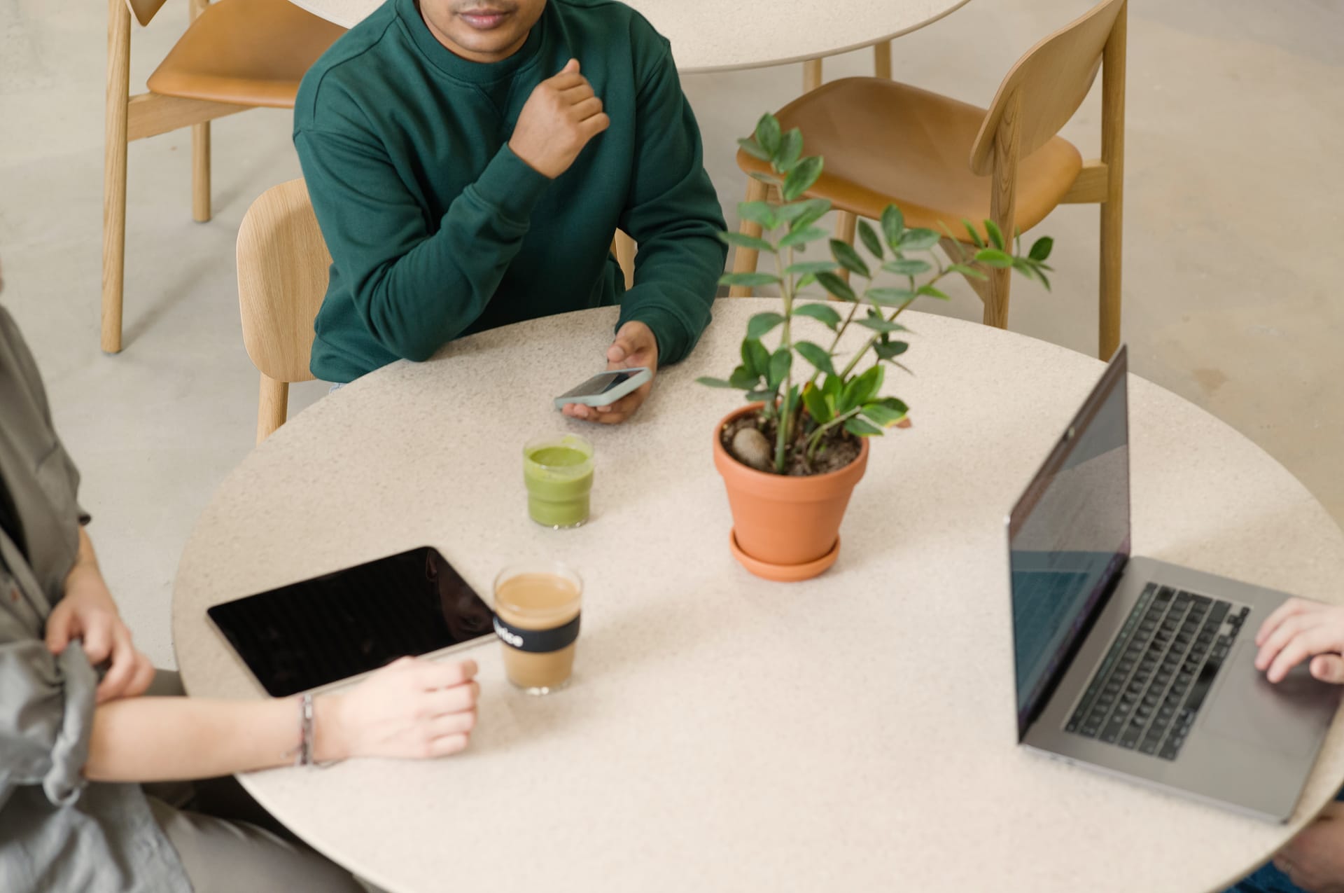 Person in green jumper at round table with laptop, tablet, potted plant and drinks during casual meeting.