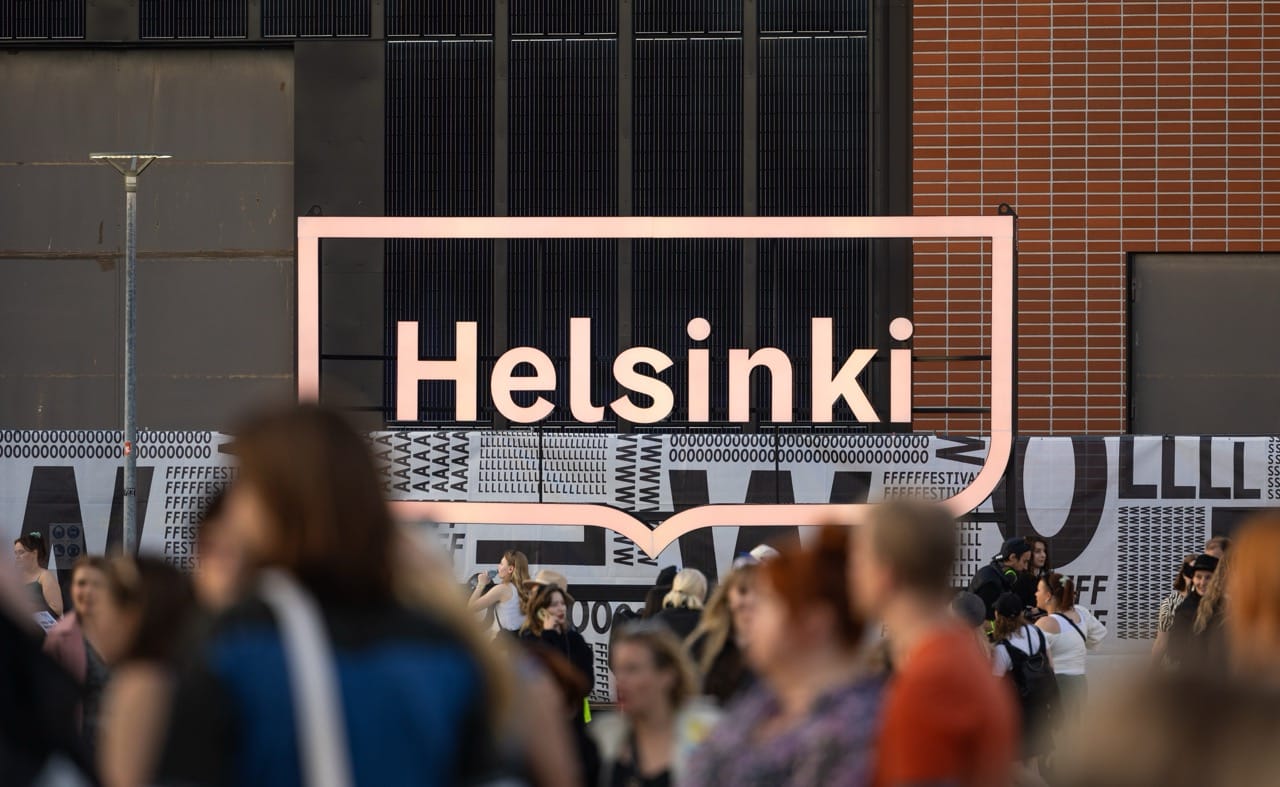 A crowd of people gathered in front of a large pink illuminated Helsinki sign on a building facade.