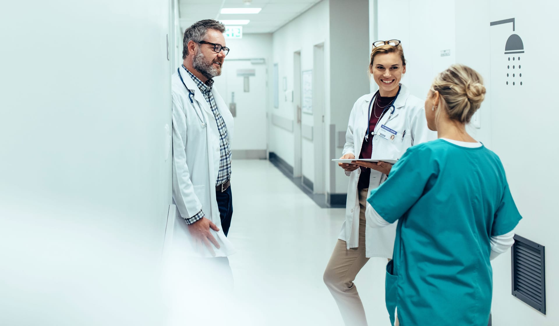 Medical professionals in white coats conversing with a nurse in teal scrubs in a bright hospital corridor.