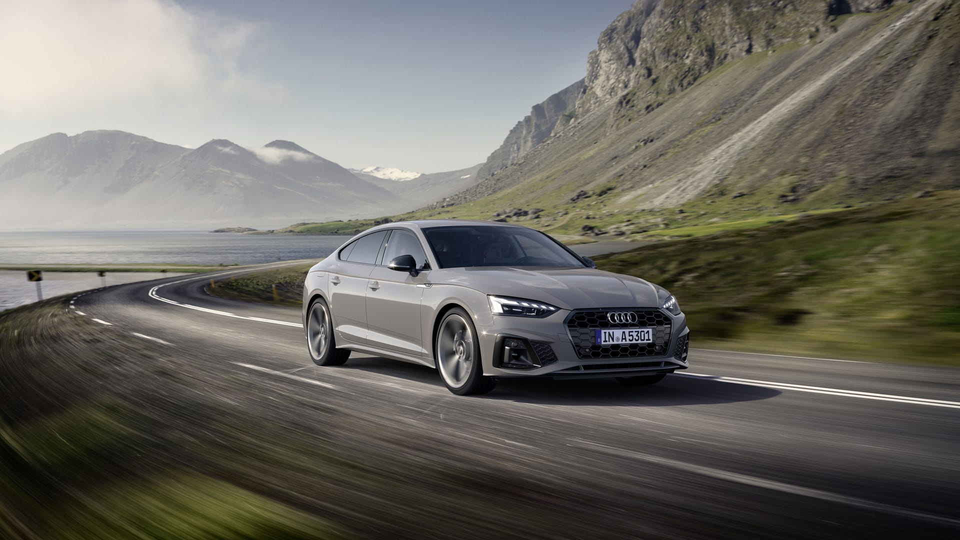 Silver Audi A5 driving on coastal mountain road with dramatic rocky cliffs and water views under clear sky.