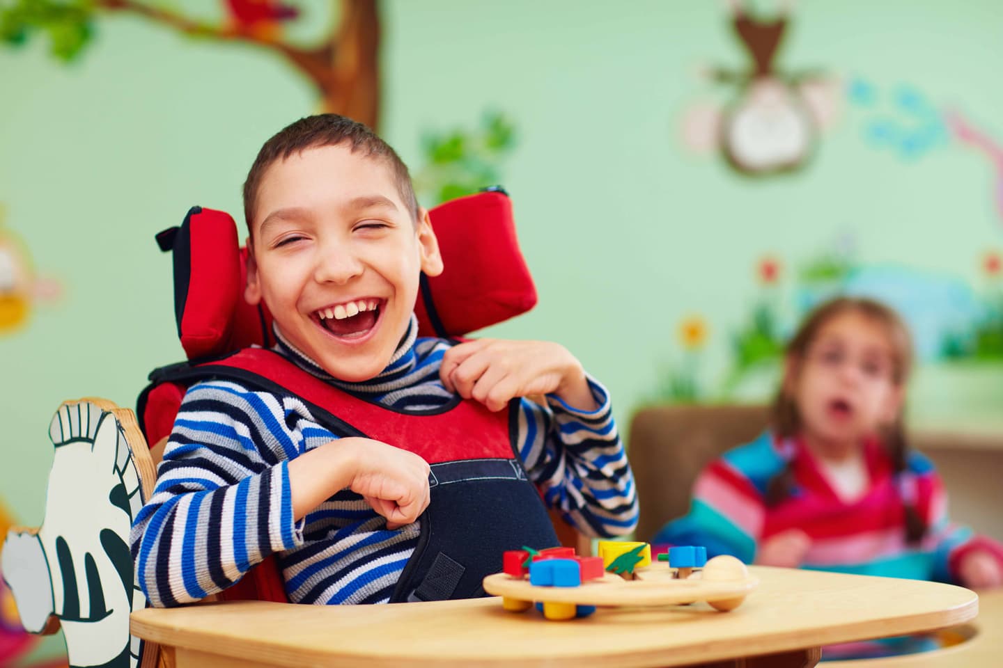 Child in wheelchair laughing joyfully with colourful toys on table in classroom with painted walls