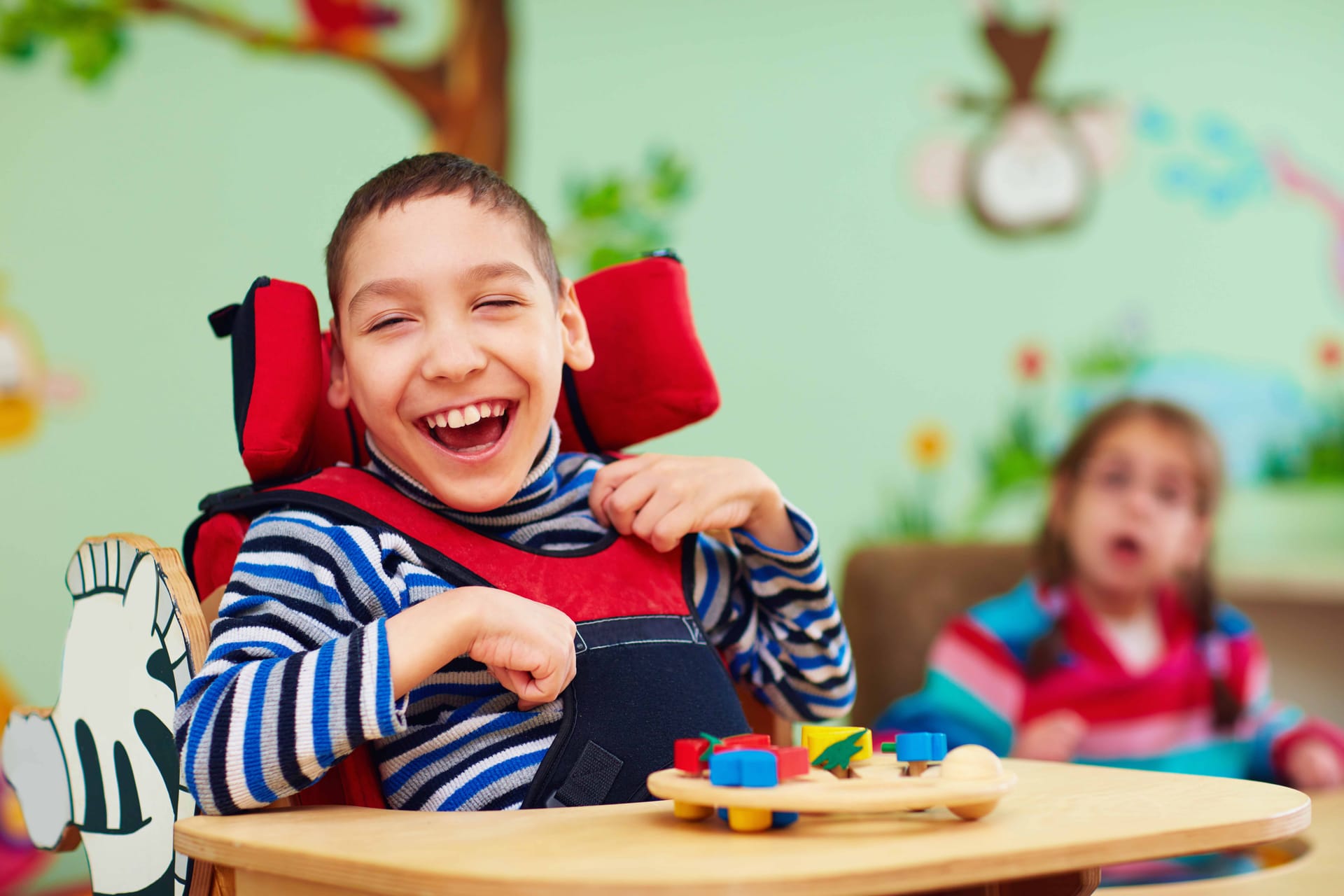Child in wheelchair laughing joyfully with colourful toys on table in classroom with painted walls