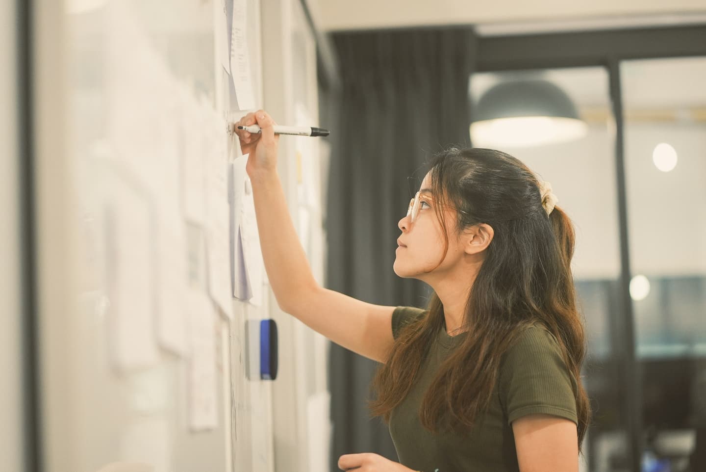 Person in green top writing on whiteboard in modern office space, focused on their work.