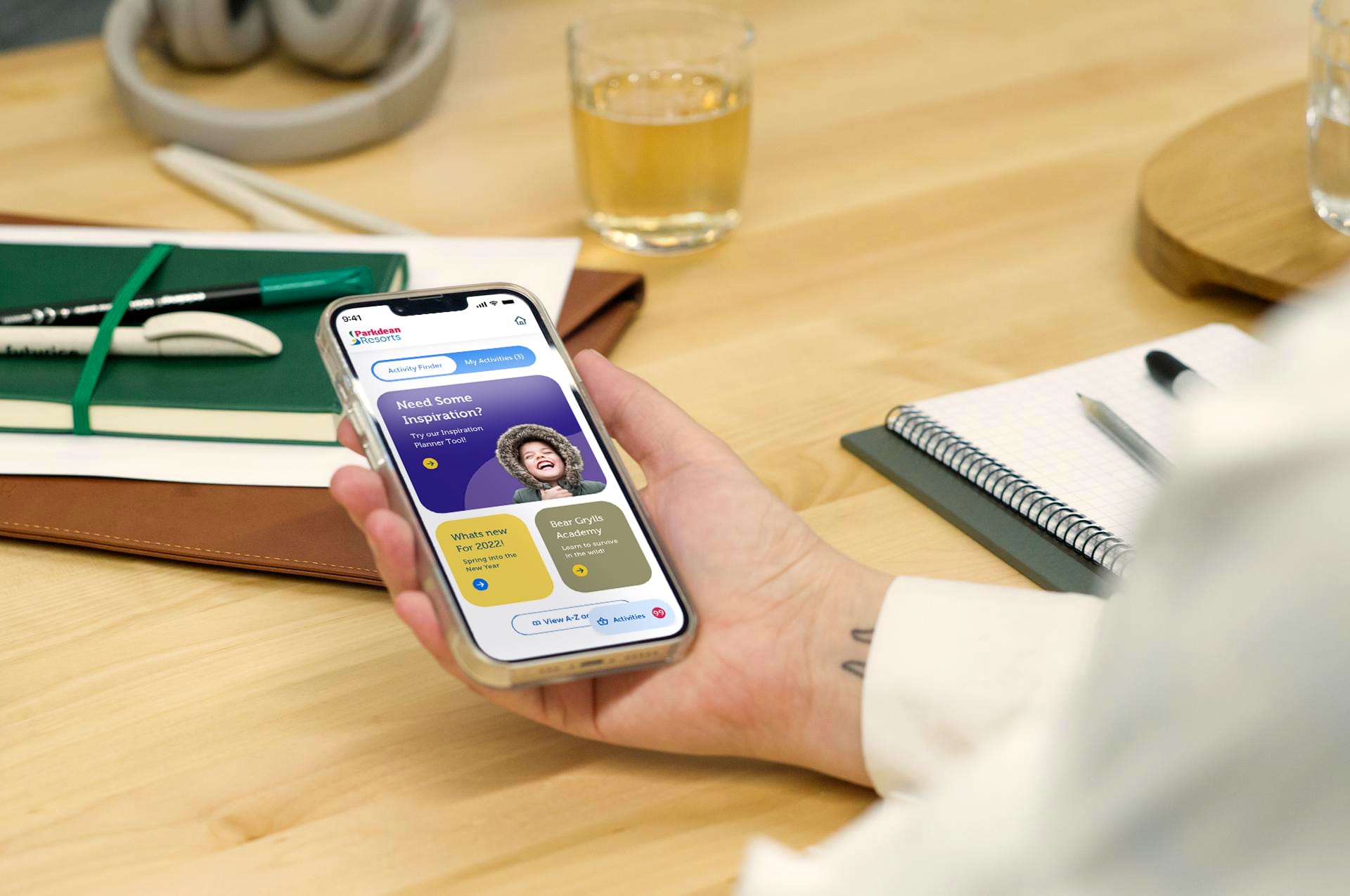 Hand holding smartphone displaying Davidson Resorts app with activity finder screen, on wooden desk with notebooks and drink.