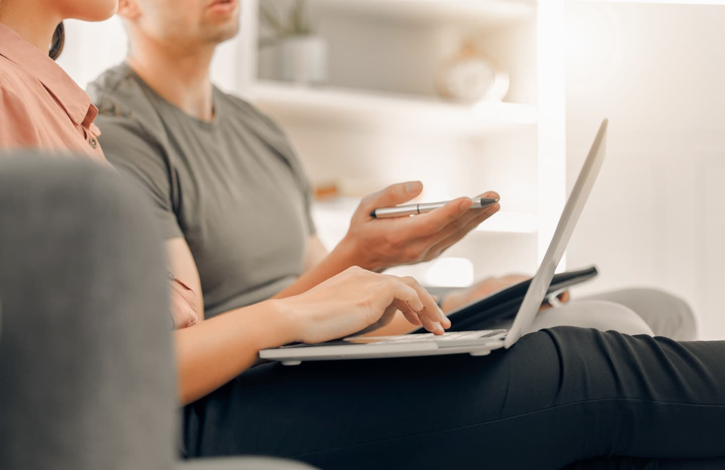 Two people sitting closely, one using a laptop while gesturing with a pen, in a bright, minimalist home setting.