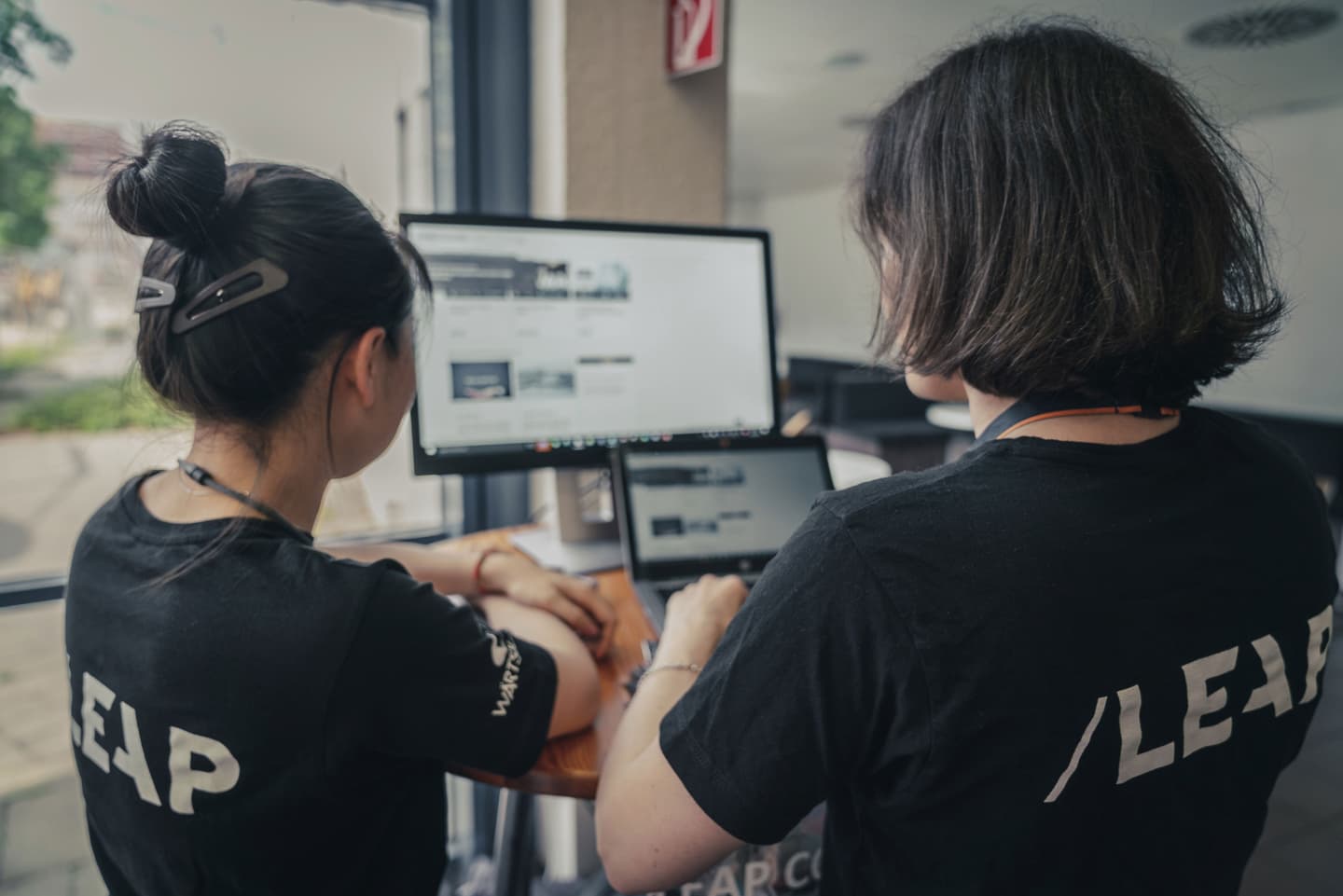 Two colleagues in black t-shirts collaborating at a workstation with multiple computer monitors in an office setting.