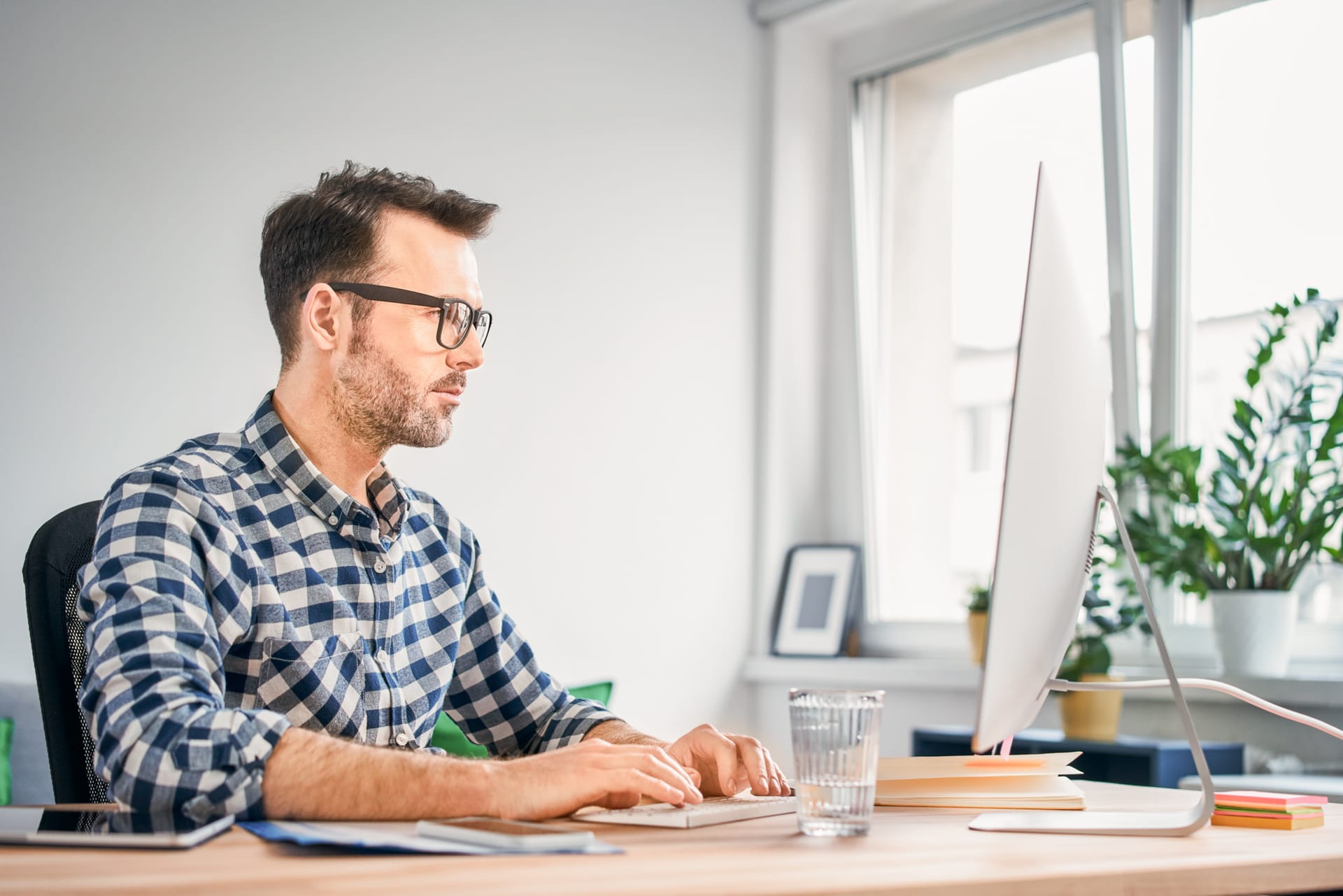 Person in checked shirt working at computer in bright home office with plants on windowsill.