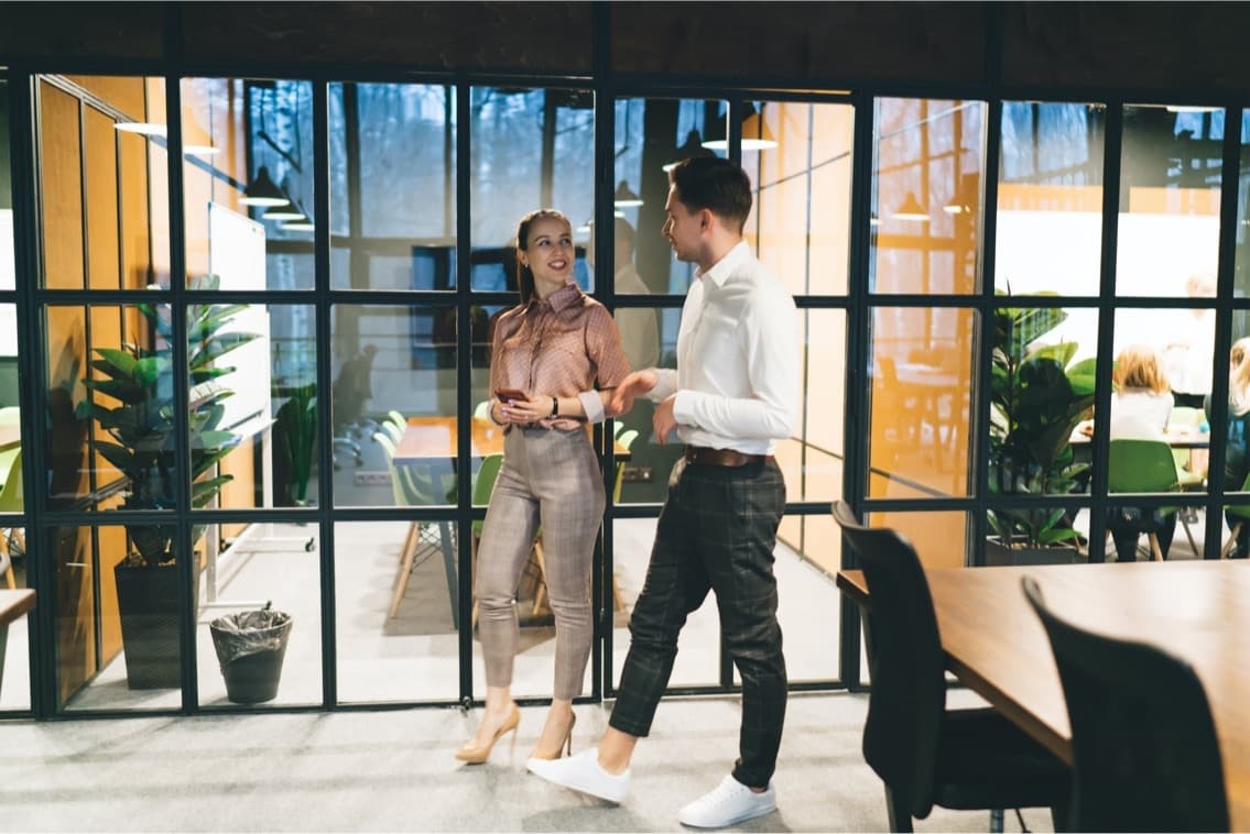 Two colleagues conversing in a modern office with glass partitions, plants, and warm lighting.