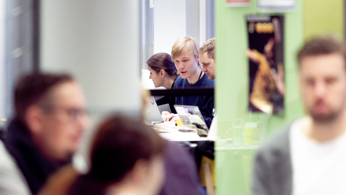 Students working together at laptops in a classroom with green walls, viewed through a doorway.