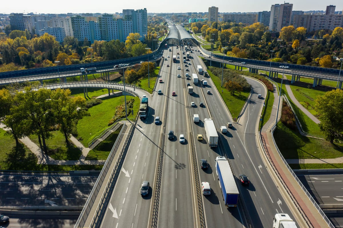 Aerial view of a busy motorway with traffic running through a city, surrounded by autumn trees and high-rise buildings.