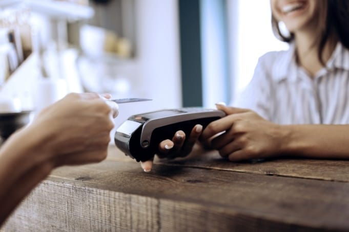 Person making contactless payment with card on payment terminal at wooden counter in bright café setting.