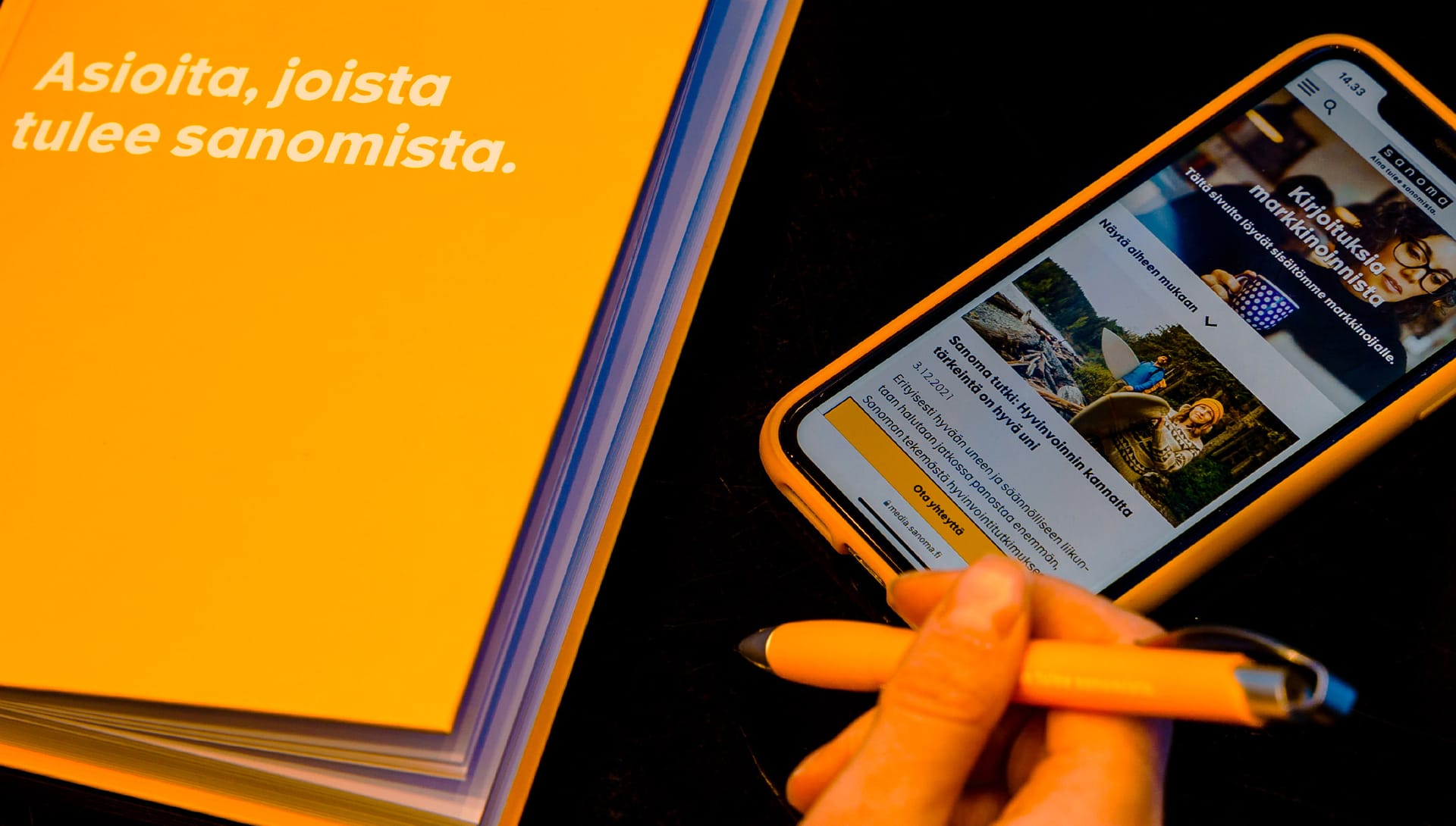 Hand holding smartphone displaying Finnish news website next to orange book with Finnish text about communications.
