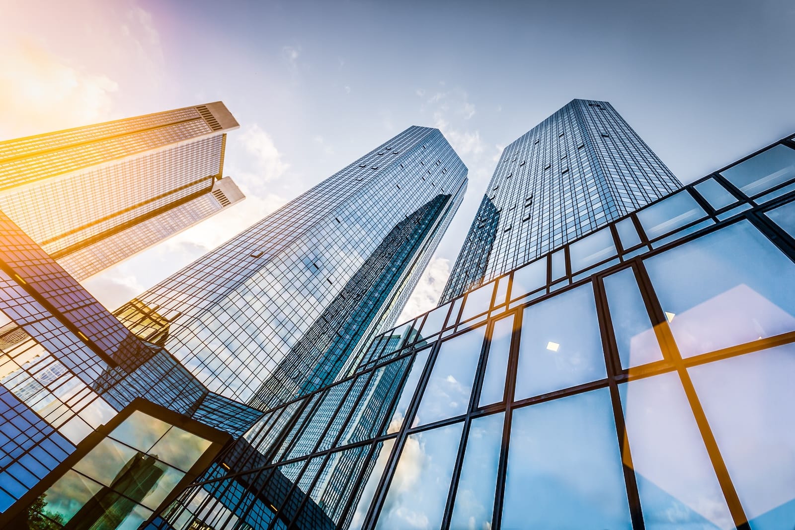 Modern glass skyscrapers viewed from below with sunlight reflecting off their facades against a blue sky.